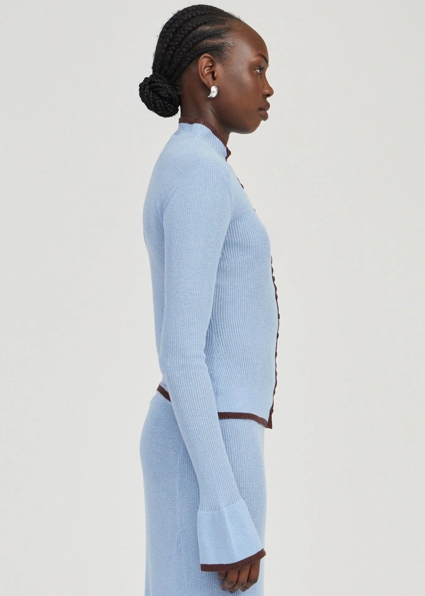 A woman shown in side profile wearing a light blue ribbed knit long-sleeve cardigan and matching skirt with dark brown trim details, against a plain light gray studio background.
