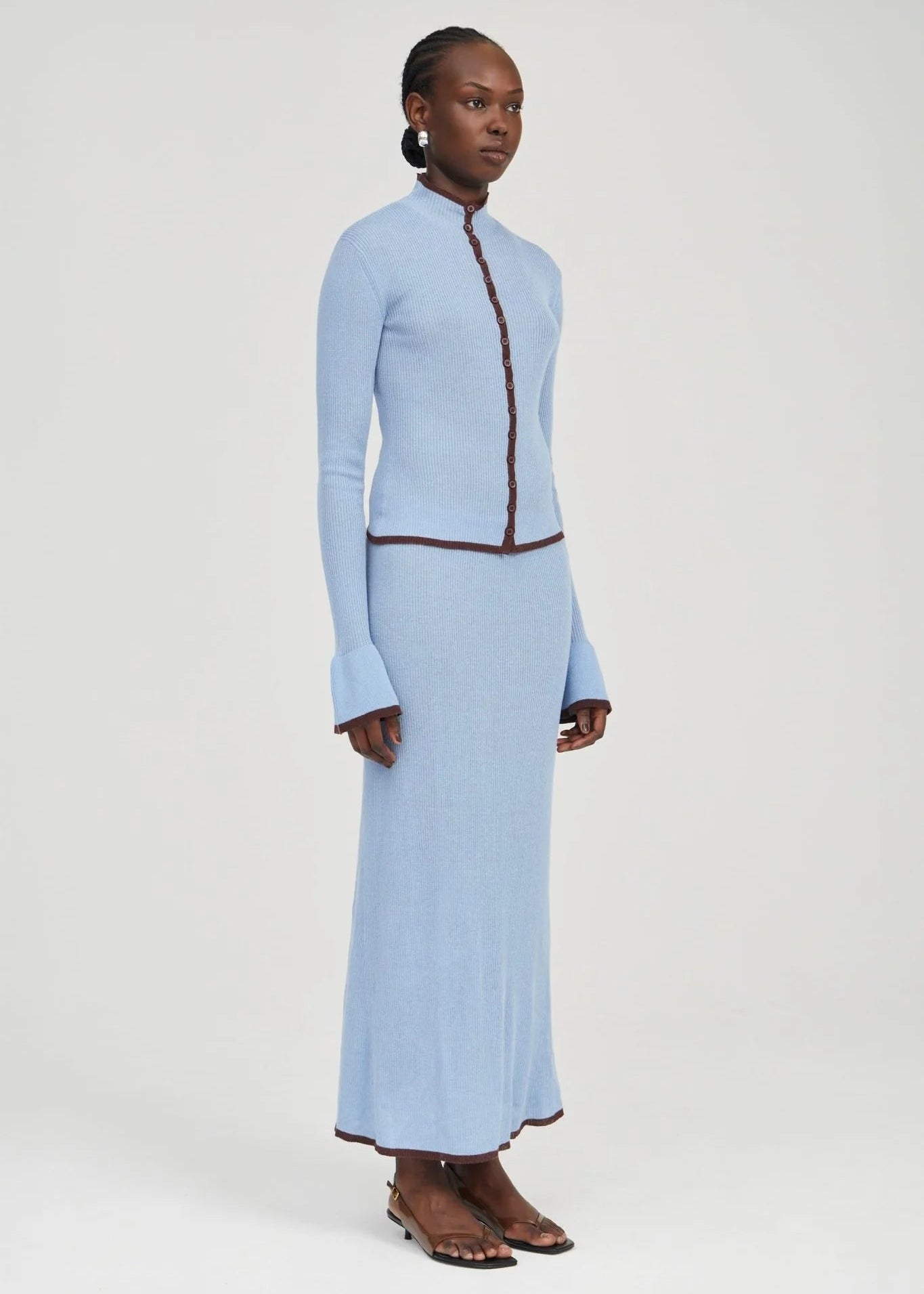 A model stands in a studio wearing a light blue knit two-piece set consisting of a long-sleeve cropped jacket with dark brown trim and a matching long skirt. She is wearing brown strappy flat sandals against a plain light gray background.