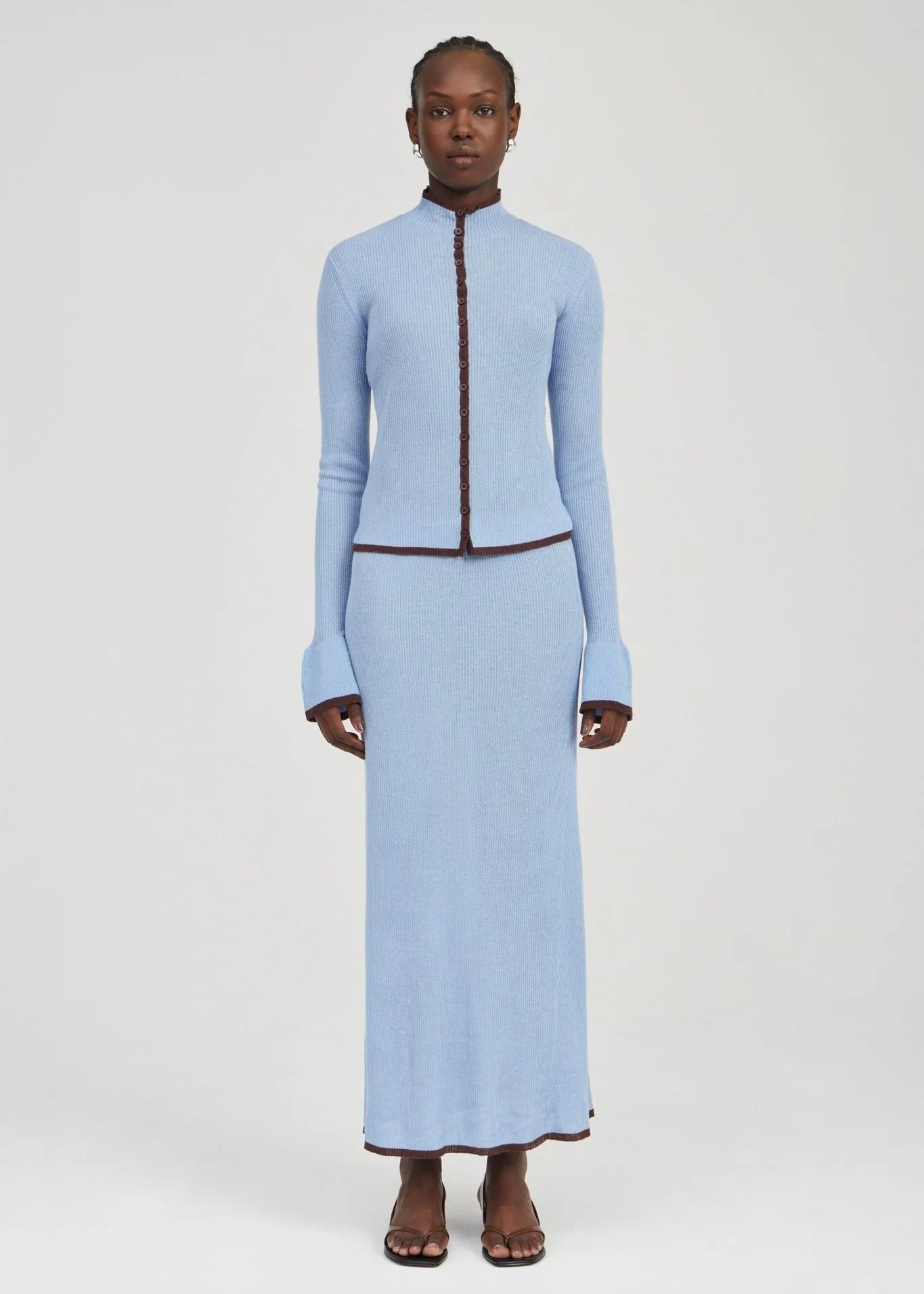 A model stands against a light gray studio background wearing a light blue knit two-piece set consisting of a cropped long-sleeve top with a dark brown trim and a matching long skirt.