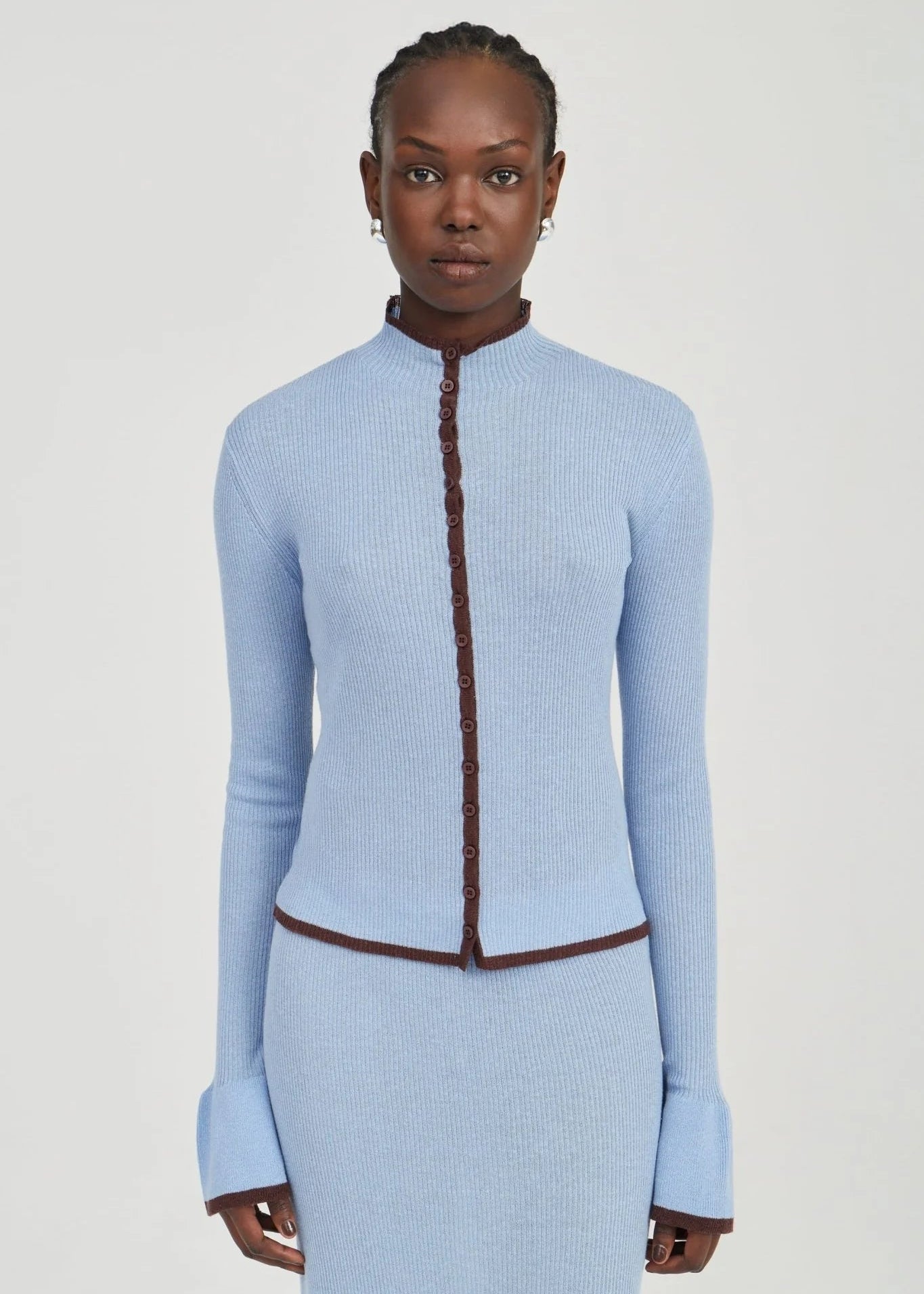 A model wearing a light blue ribbed knit two-piece set consisting of a long-sleeve cropped cardigan with a brown braided trim detail and a matching ribbed skirt, against a plain light gray studio background.