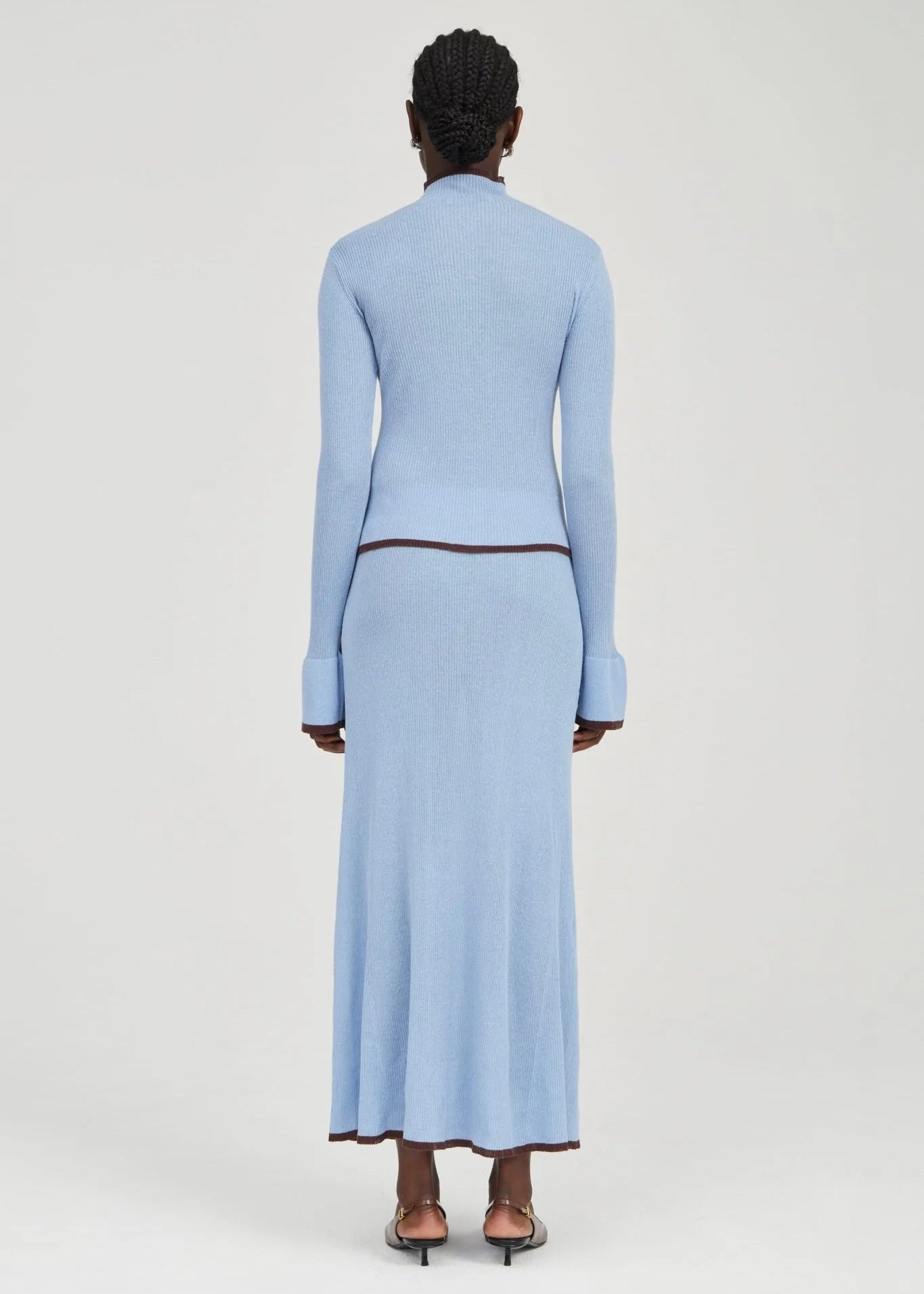 A model viewed from the back wearing a light blue knit long-sleeve top and matching midi skirt with dark brown trim, set against a plain light gray studio background.