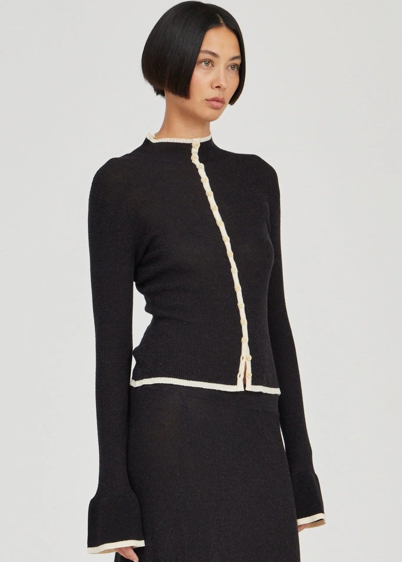 A model with a short dark bob haircut wearing a black knit long-sleeve top with cream-colored trim details, photographed against a solid light gray background.