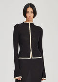 A model wearing a black knit long-sleeve top with cream-colored trim details on the collar, front placket, hem, and cuffs, paired with a matching black skirt against a light gray background.