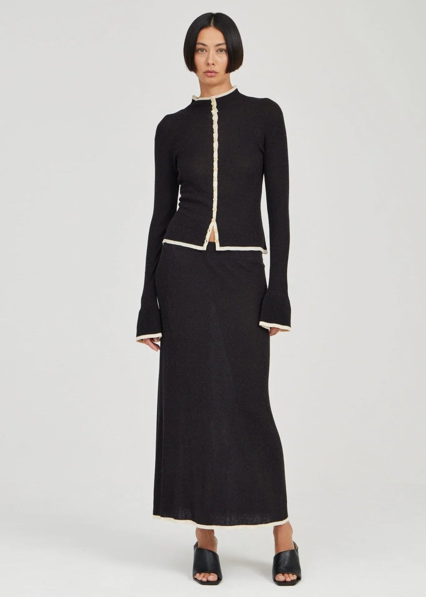 A model wearing a black long-sleeve knit top and matching maxi skirt set with cream-colored trim details, standing against a light gray studio background.