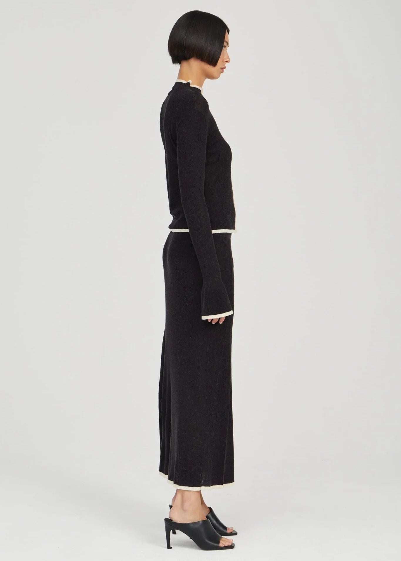 A model shown from the side profile wearing a black ribbed knit long-sleeve top and matching midi skirt with thin white contrast trim at the waist and hem, paired with black mule heels, against a light gray studio background.