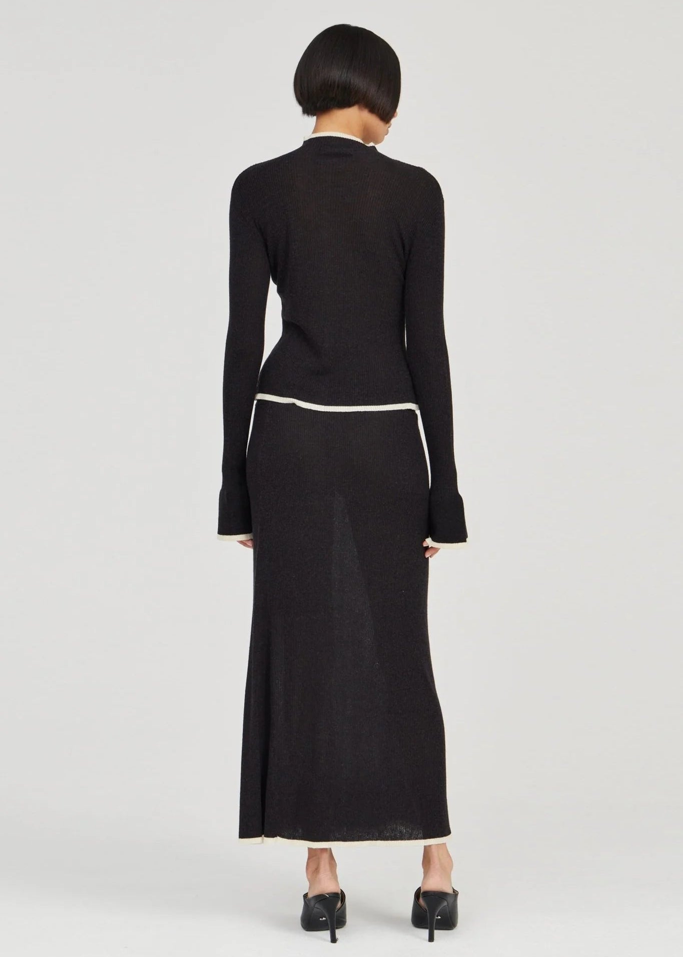 A model viewed from the back wearing a black long-sleeve knit top and matching maxi skirt with a thin white waistband, paired with black pointed-toe heels, against a light gray studio background.