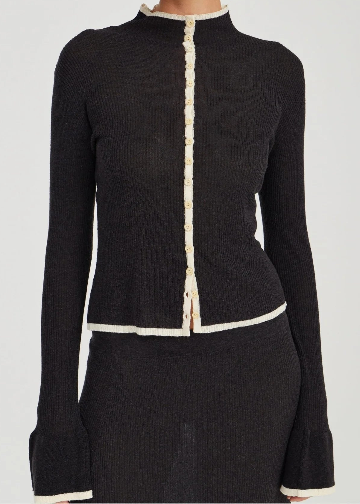 A model wearing a black ribbed knit long-sleeve top with cream-colored trim detailing the collar, front placket, hem, and cuffs, paired with a matching black skirt.