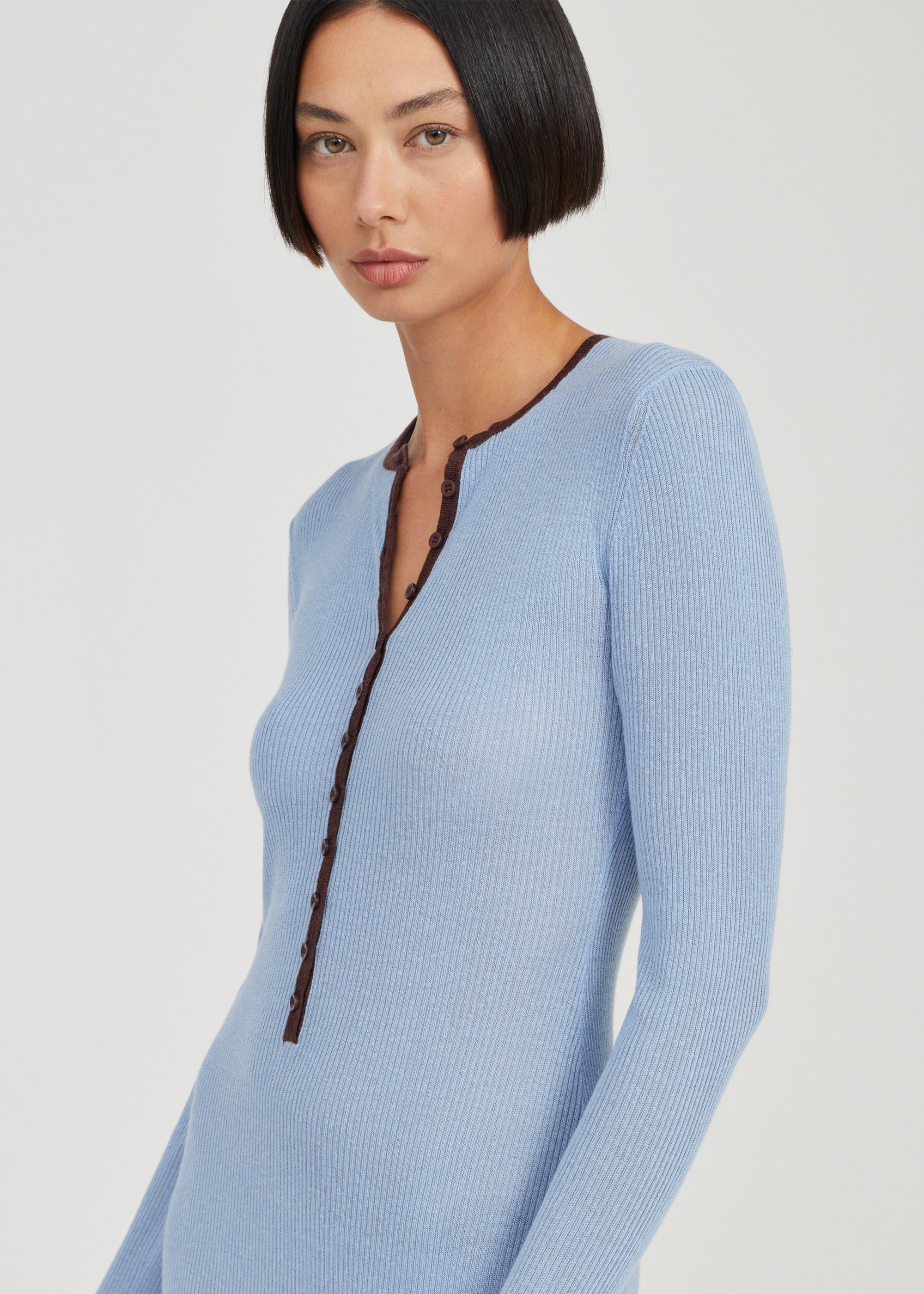 A woman wearing a light blue ribbed knit long-sleeve top with a dark brown braided trim detail along the split neckline, shown against a plain light gray studio background.