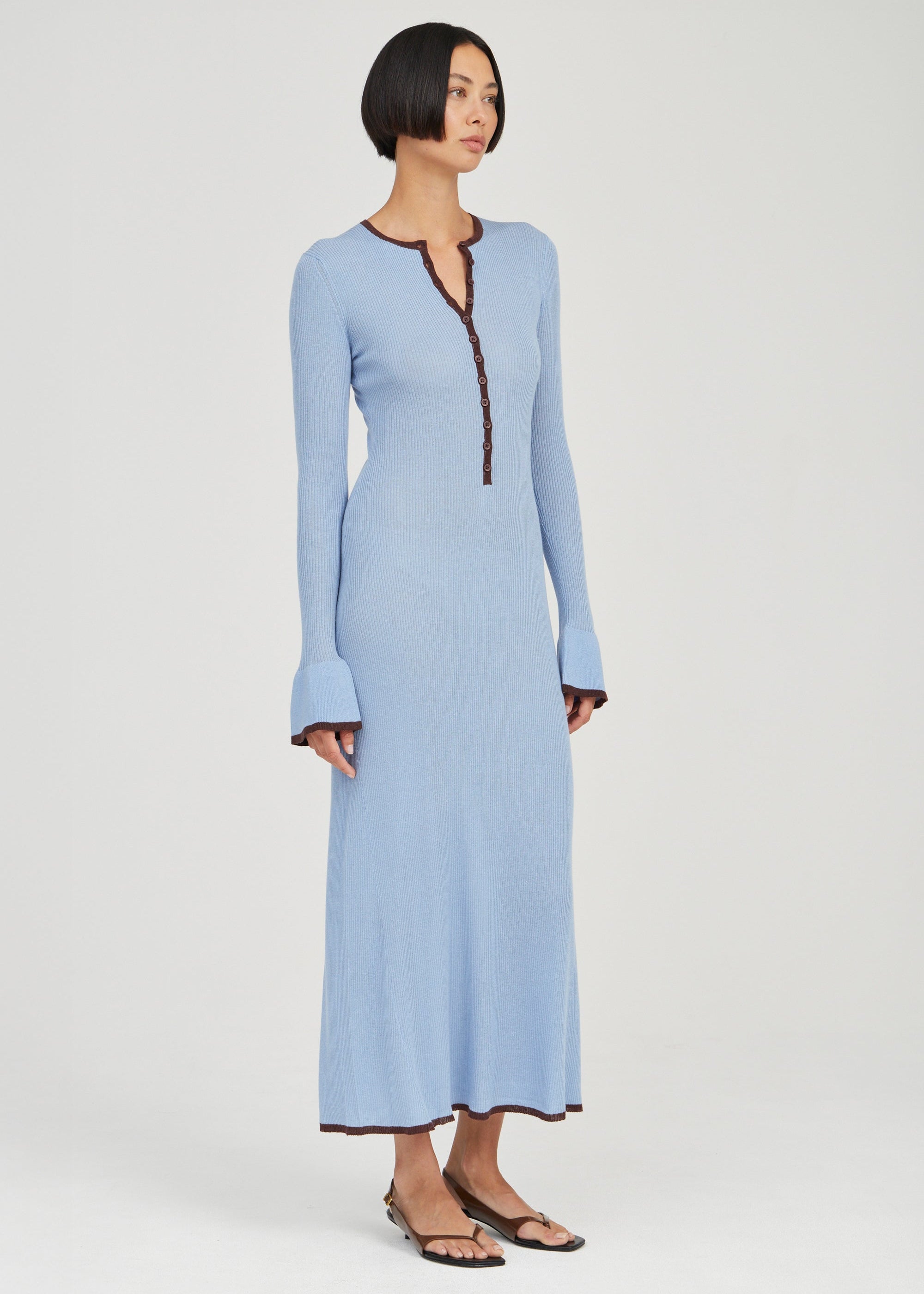 A woman modeling a light blue long-sleeve maxi dress with dark brown trim along the split neckline, cuffs, and hem, paired with black flat sandals against a light gray studio background.