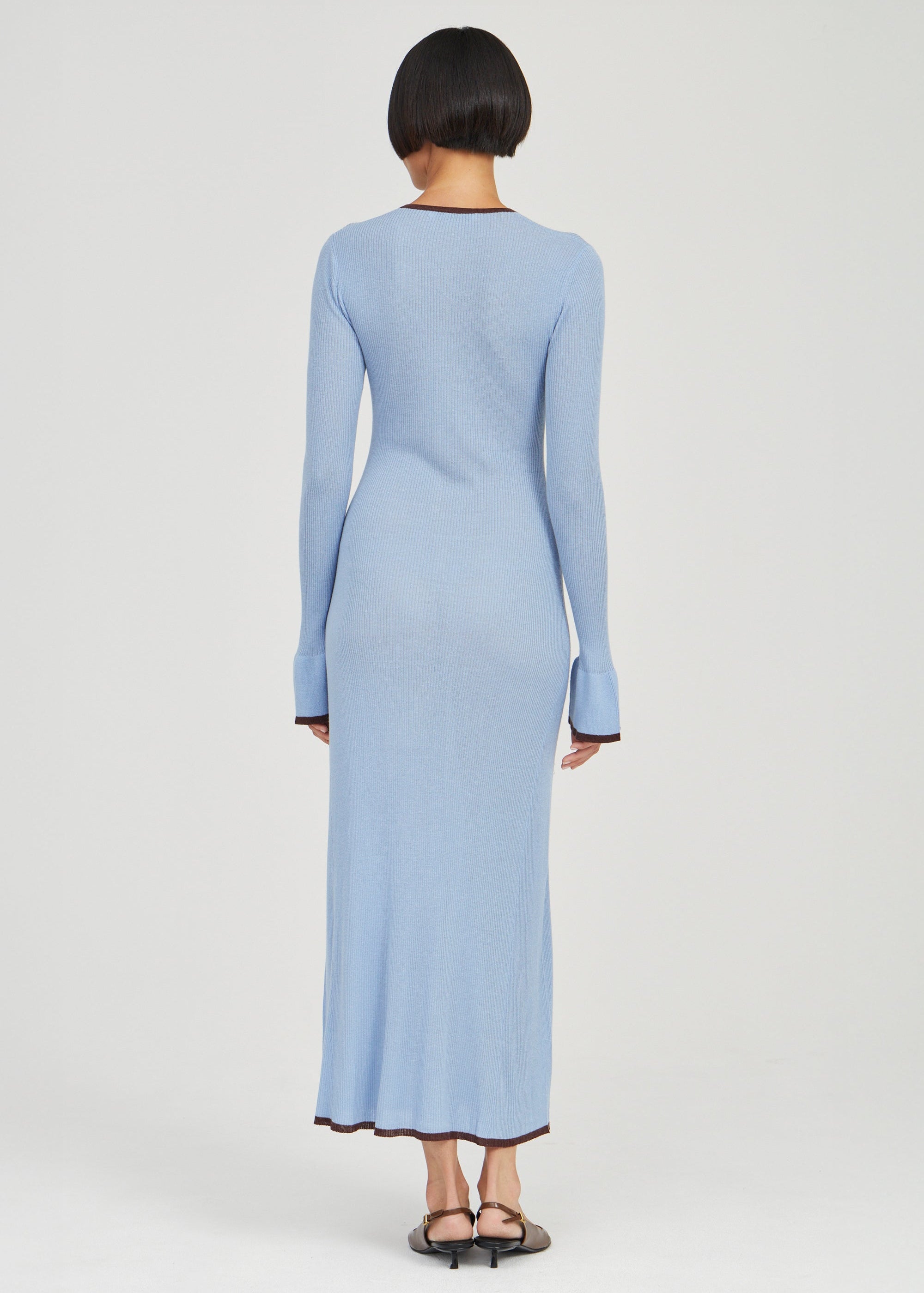 A model viewed from the back wearing a light blue long-sleeve maxi dress with dark trim at the collar, cuffs, and hem, paired with black flat sandals, against a light gray studio background.