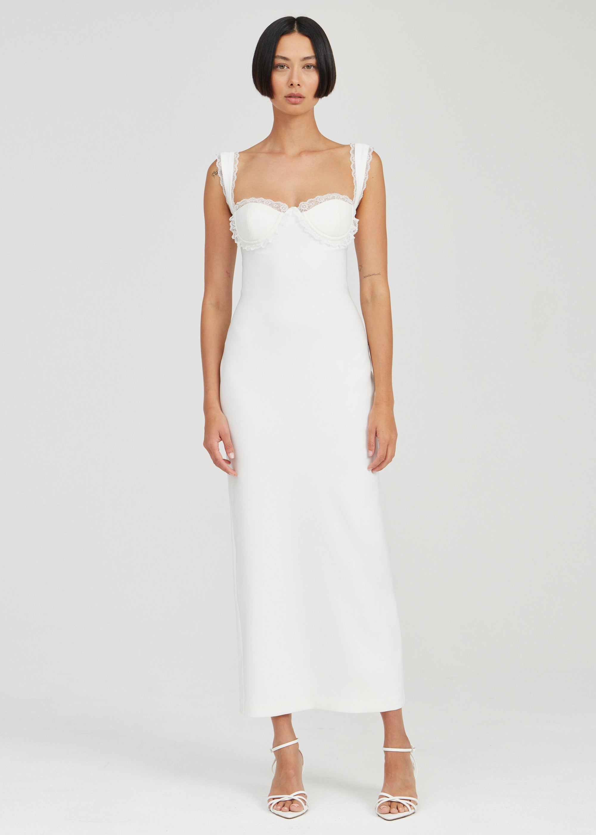 A woman modeling a white sleeveless midi dress with ruffled shoulder straps and a structured sweetheart neckline, paired with white strappy high-heeled sandals, against a light gray studio background.