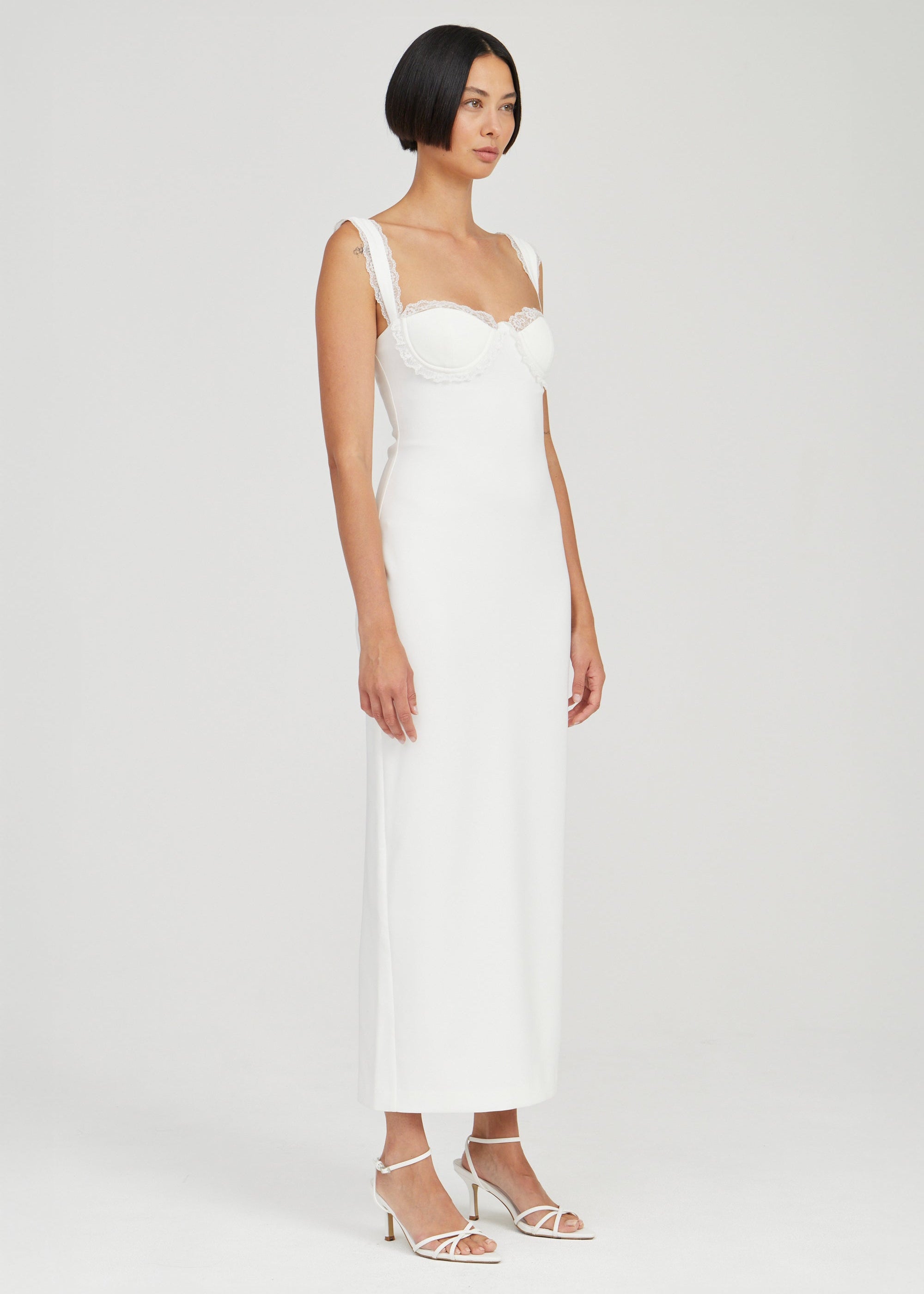 A woman modeling a white sleeveless midi dress with a sweetheart neckline and structured shoulder straps, paired with white strappy heeled sandals, against a light gray studio background.
