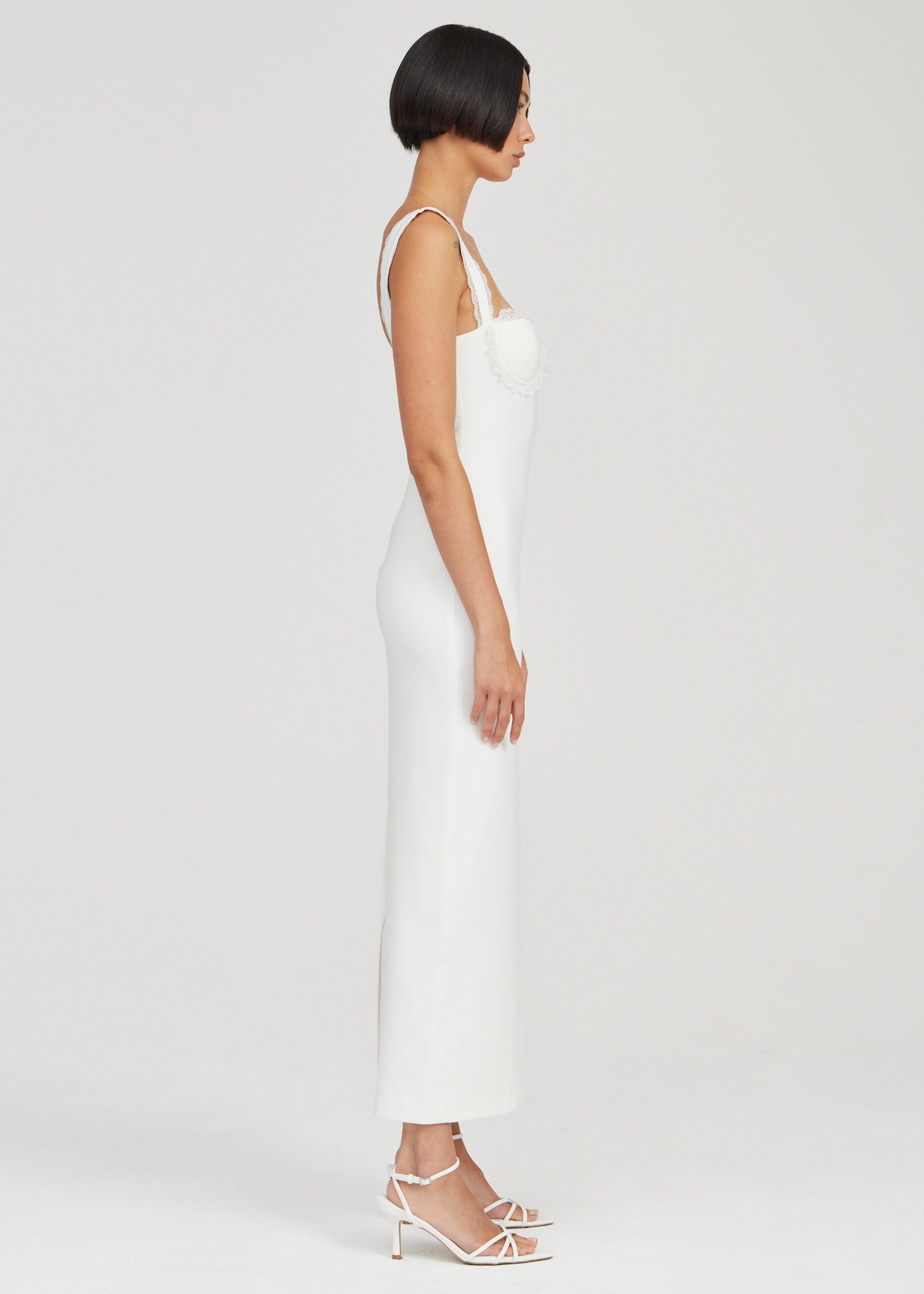 A woman shown in side profile wearing a fitted white sleeveless midi dress with thin straps and white strappy heeled sandals against a plain light gray studio background.