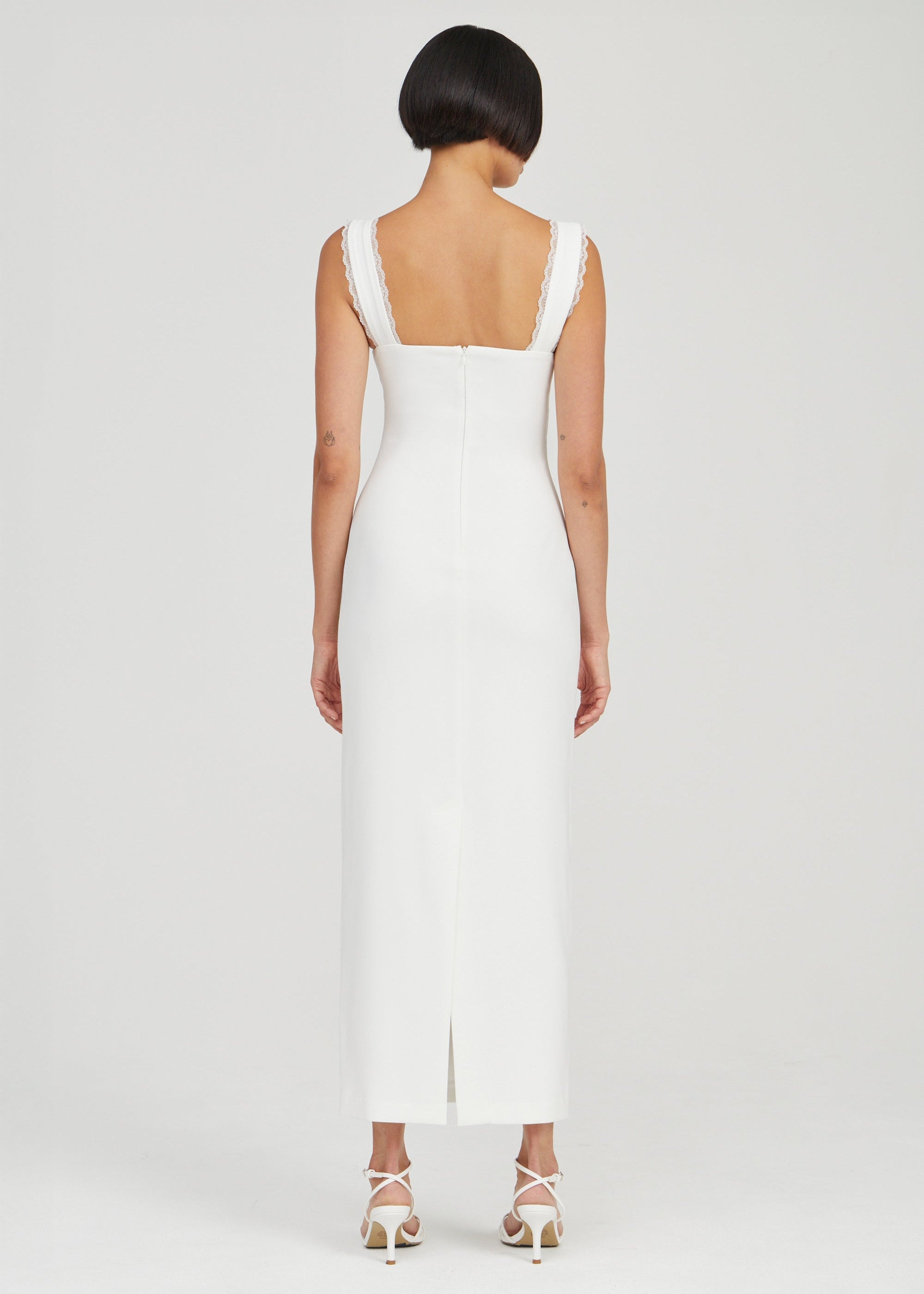 A woman viewed from the back wearing a sleeveless white maxi dress with a square neckline and a front slit, paired with white strappy high-heeled sandals, against a light gray studio background.