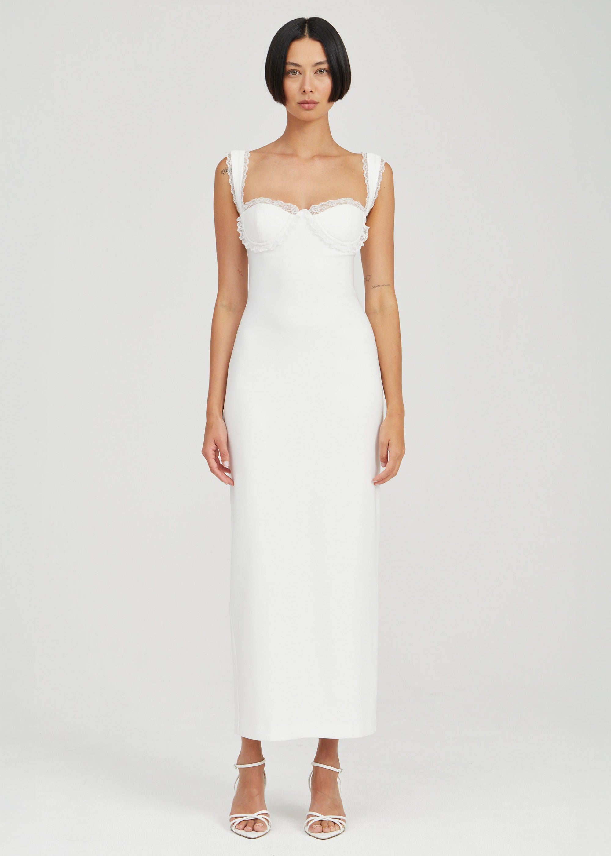 A woman modeling a white sleeveless midi dress with a structured sweetheart neckline, ruffled shoulder straps, and a fitted silhouette, photographed against a light gray studio background.