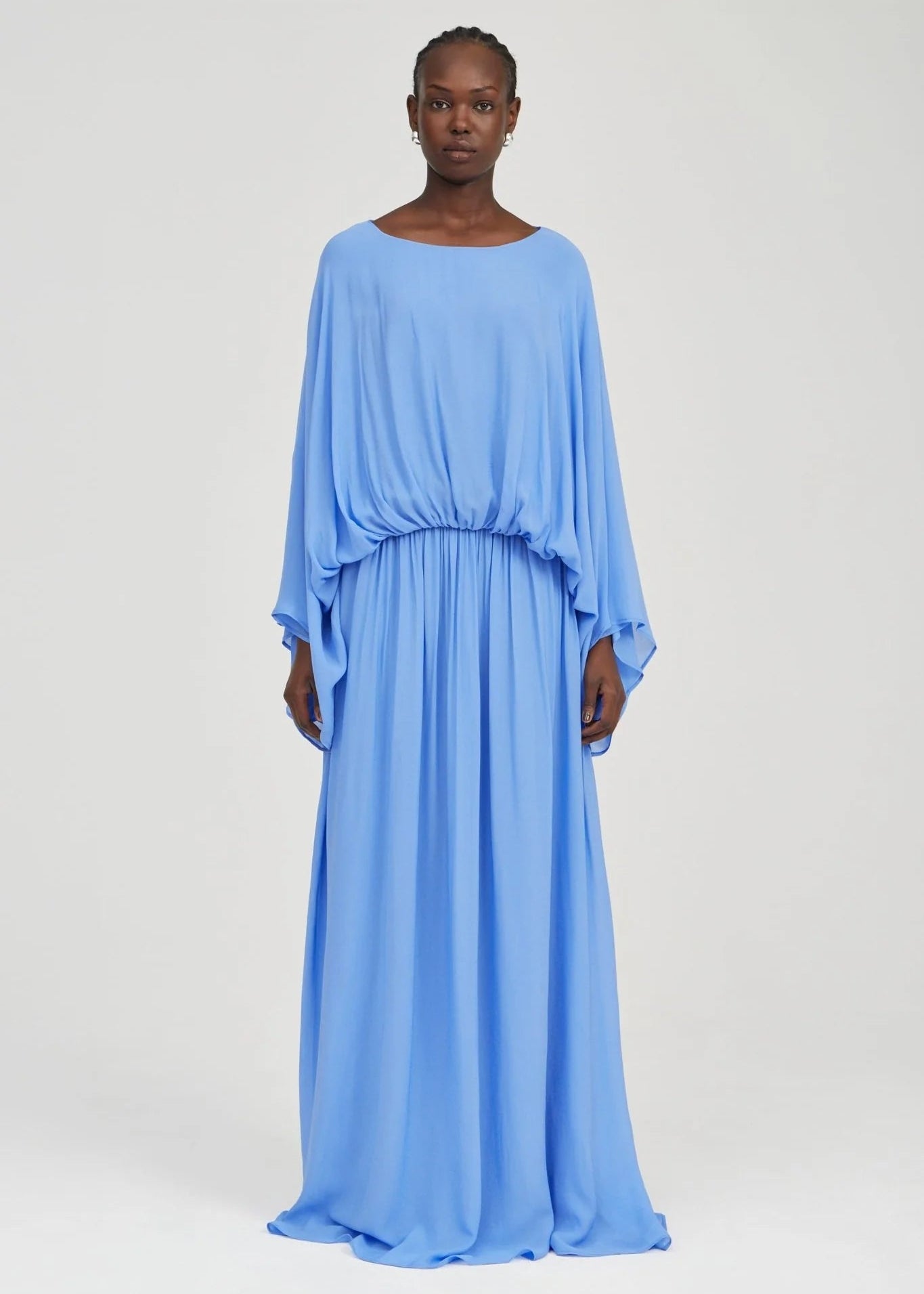 A model wearing a light blue, long-sleeved maxi dress with a gathered waist, photographed against a neutral light gray studio background.