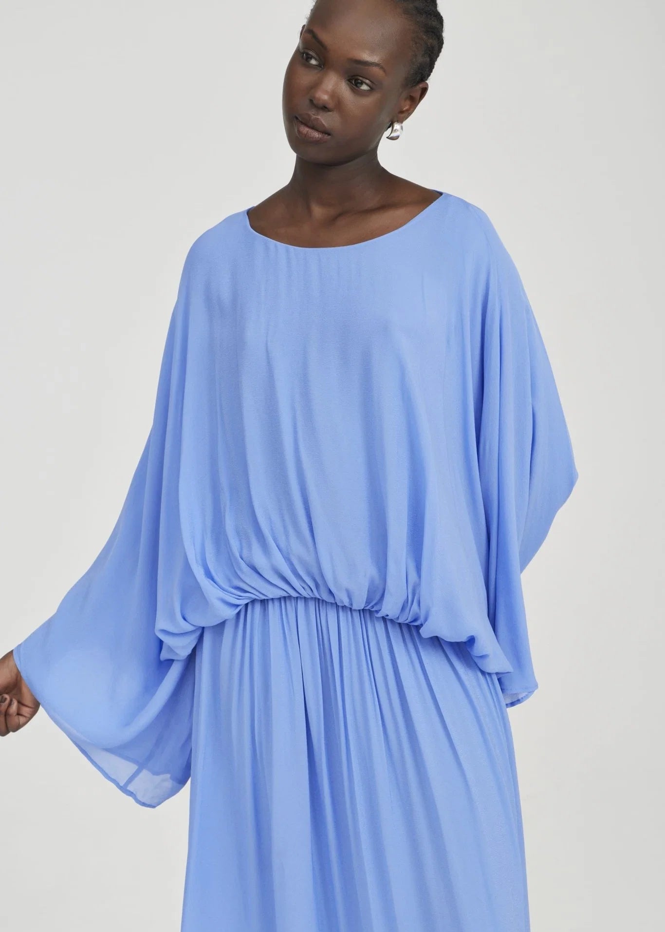 A woman wearing a light blue, flowing maxi dress with wide draped sleeves and a gathered waist, photographed against a plain light gray studio background.