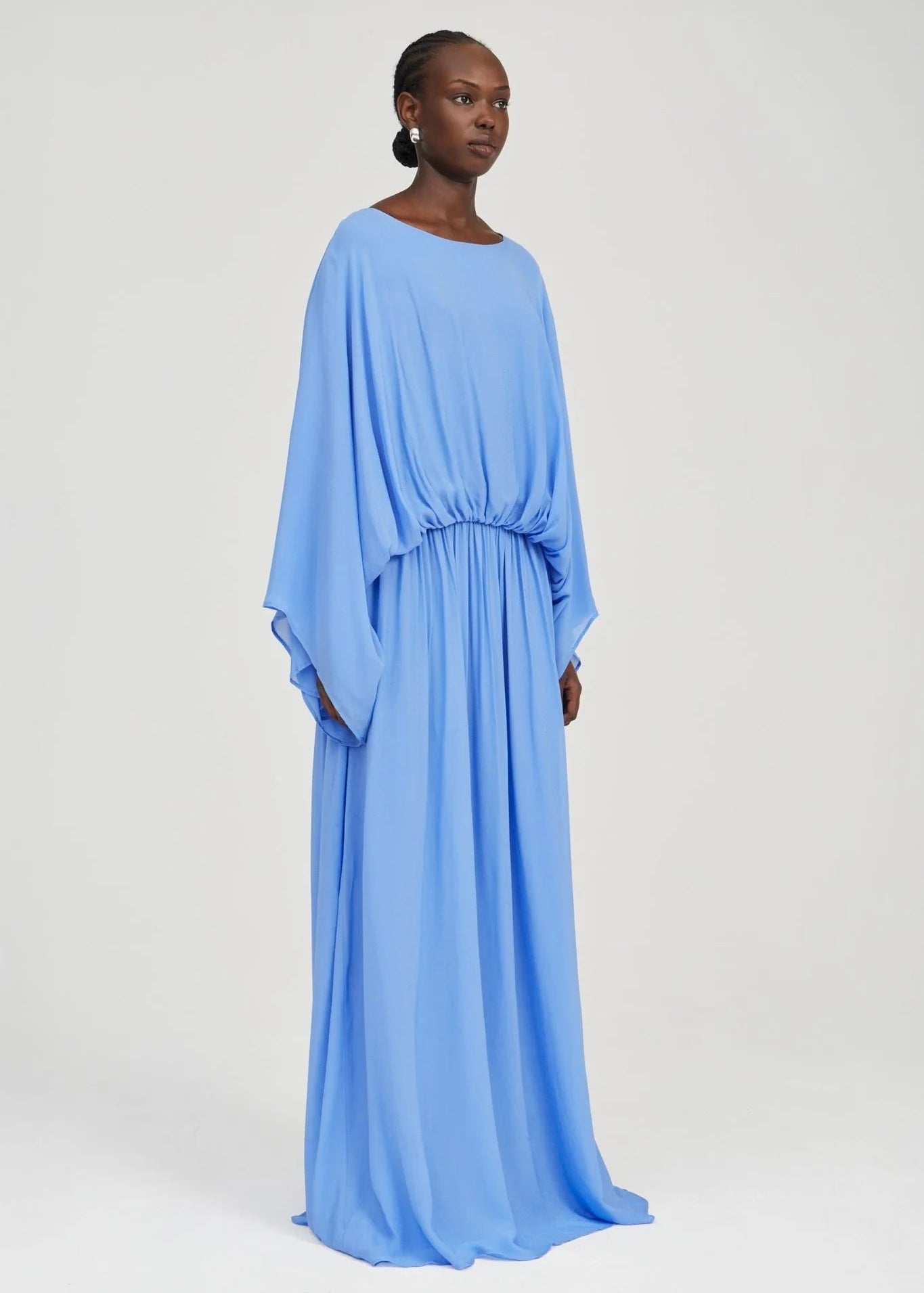 A woman modeling a flowing light blue maxi dress with wide draped sleeves and a gathered waist, photographed against a plain light gray studio background.