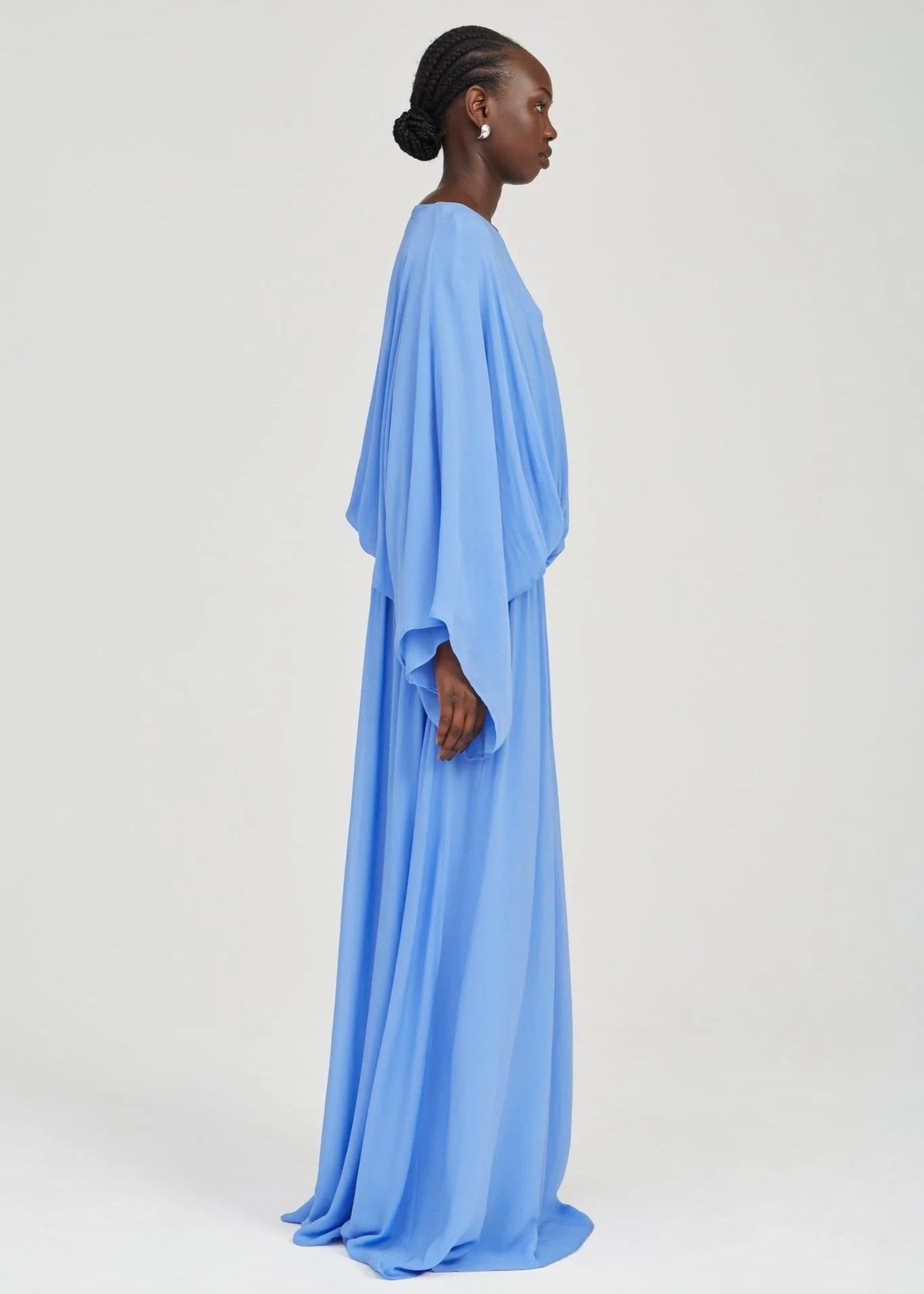 A model shown in profile wearing a flowing light blue caftan or long dress against a plain light gray studio background.