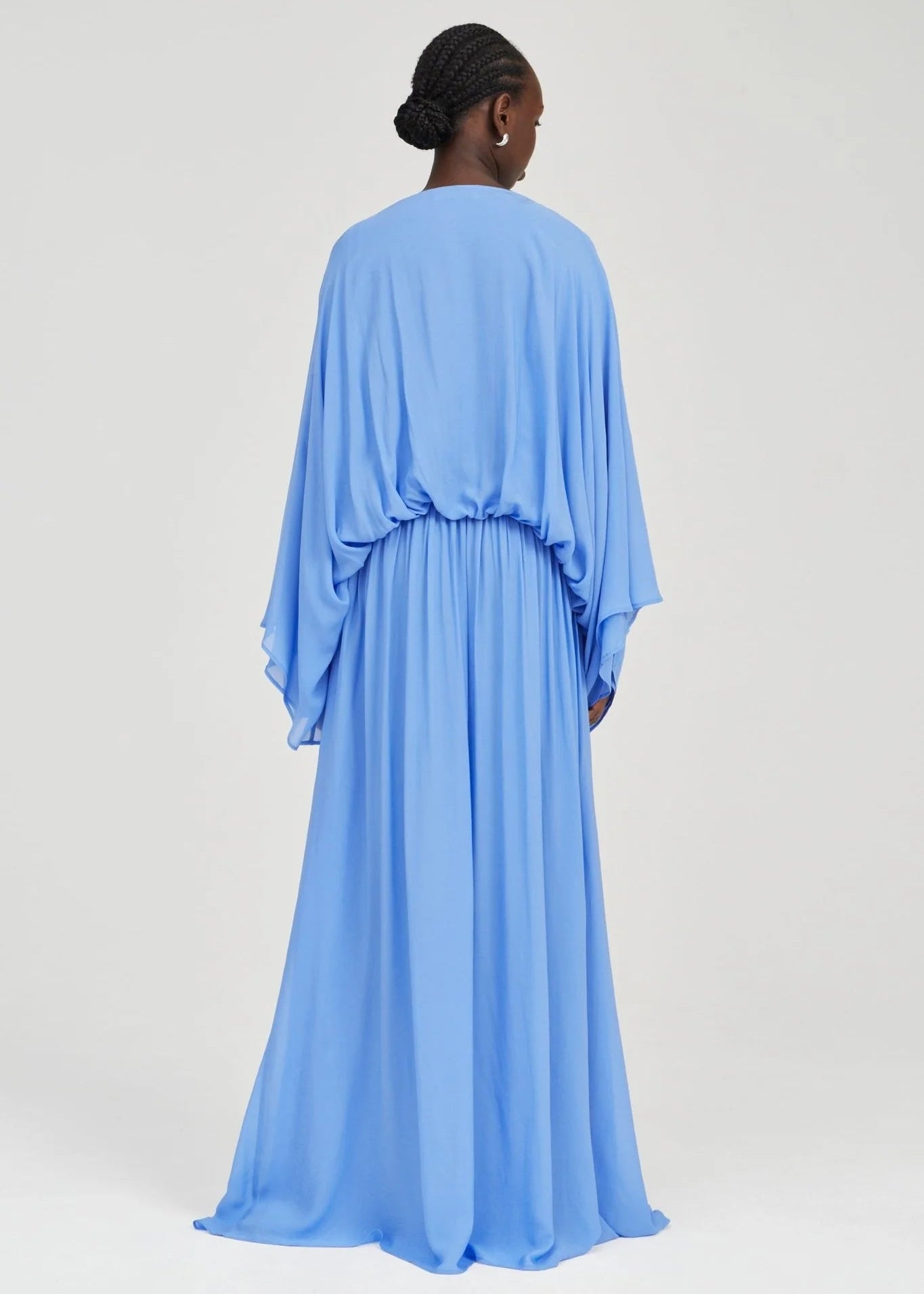 A model viewed from the back wearing a flowing light blue maxi dress with draped, wide sleeves and a gathered waist, set against a plain light gray studio background.
