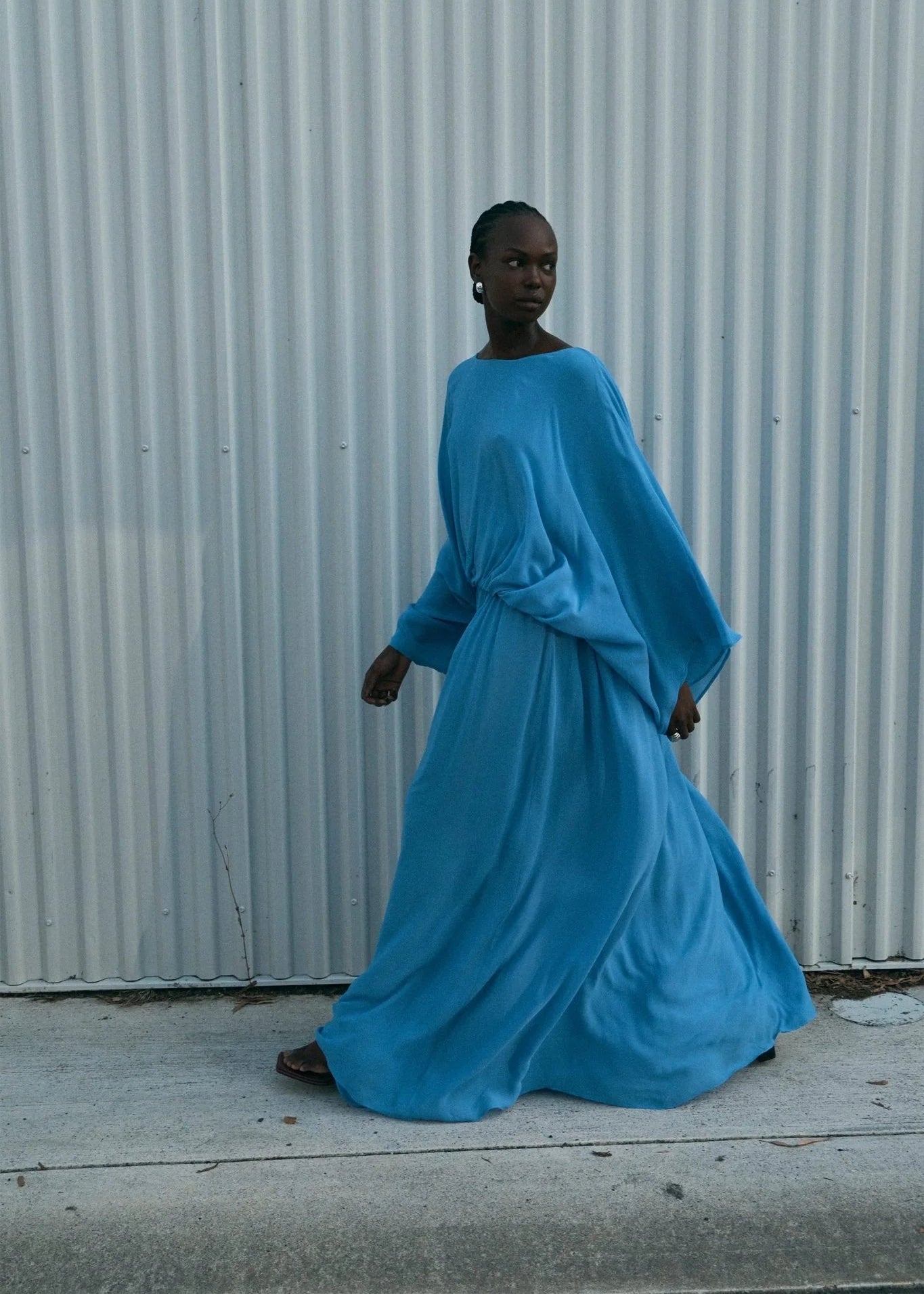 A model wearing a flowing, long-sleeved blue maxi dress walks in front of a corrugated metal wall.