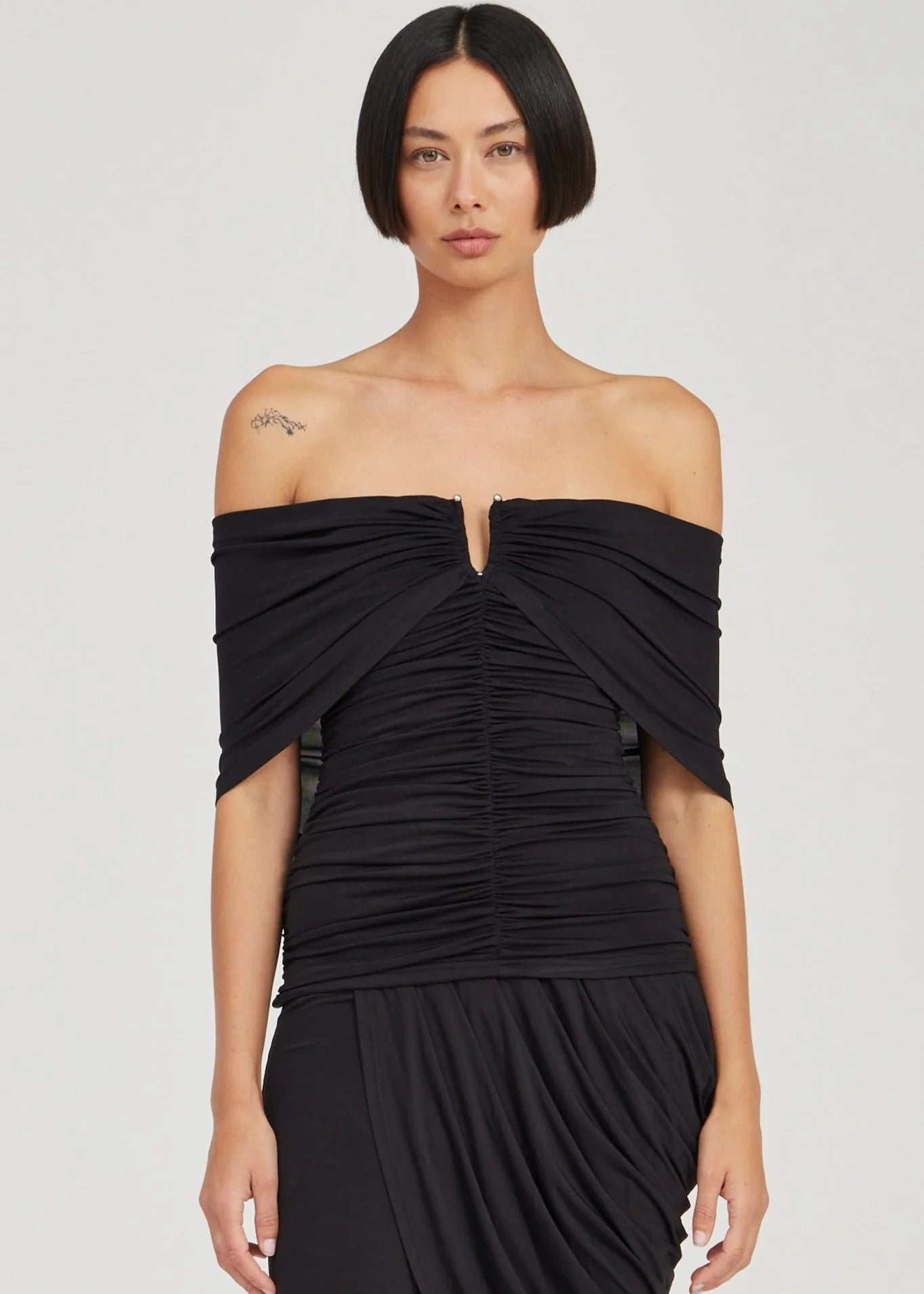 A model with a short dark bob hairstyle wearing a black off-the-shoulder ruched dress against a light gray studio background.