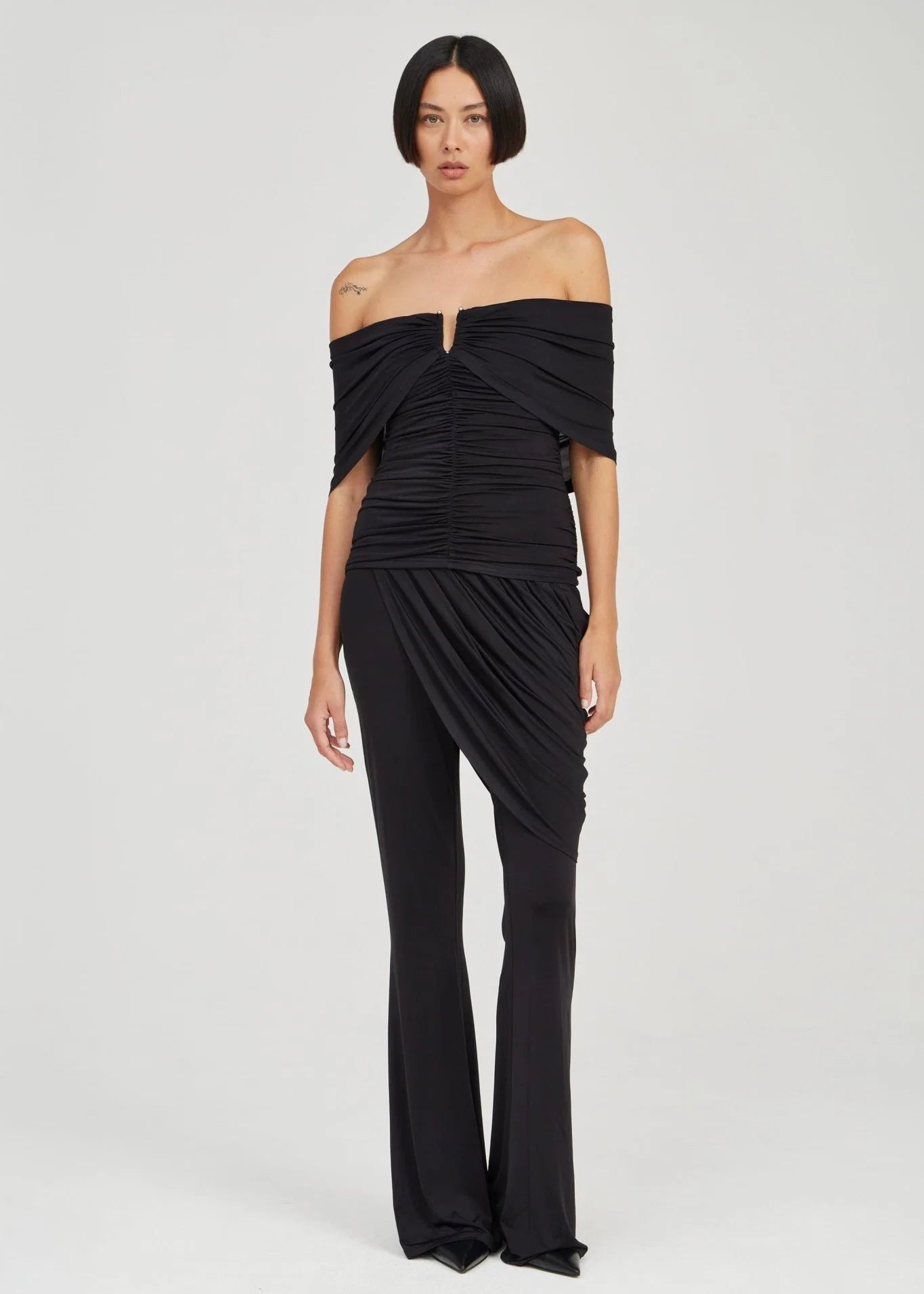 A model wearing a black off-the-shoulder draped jumpsuit with a ruched bodice and flared legs, photographed against a light gray studio background.