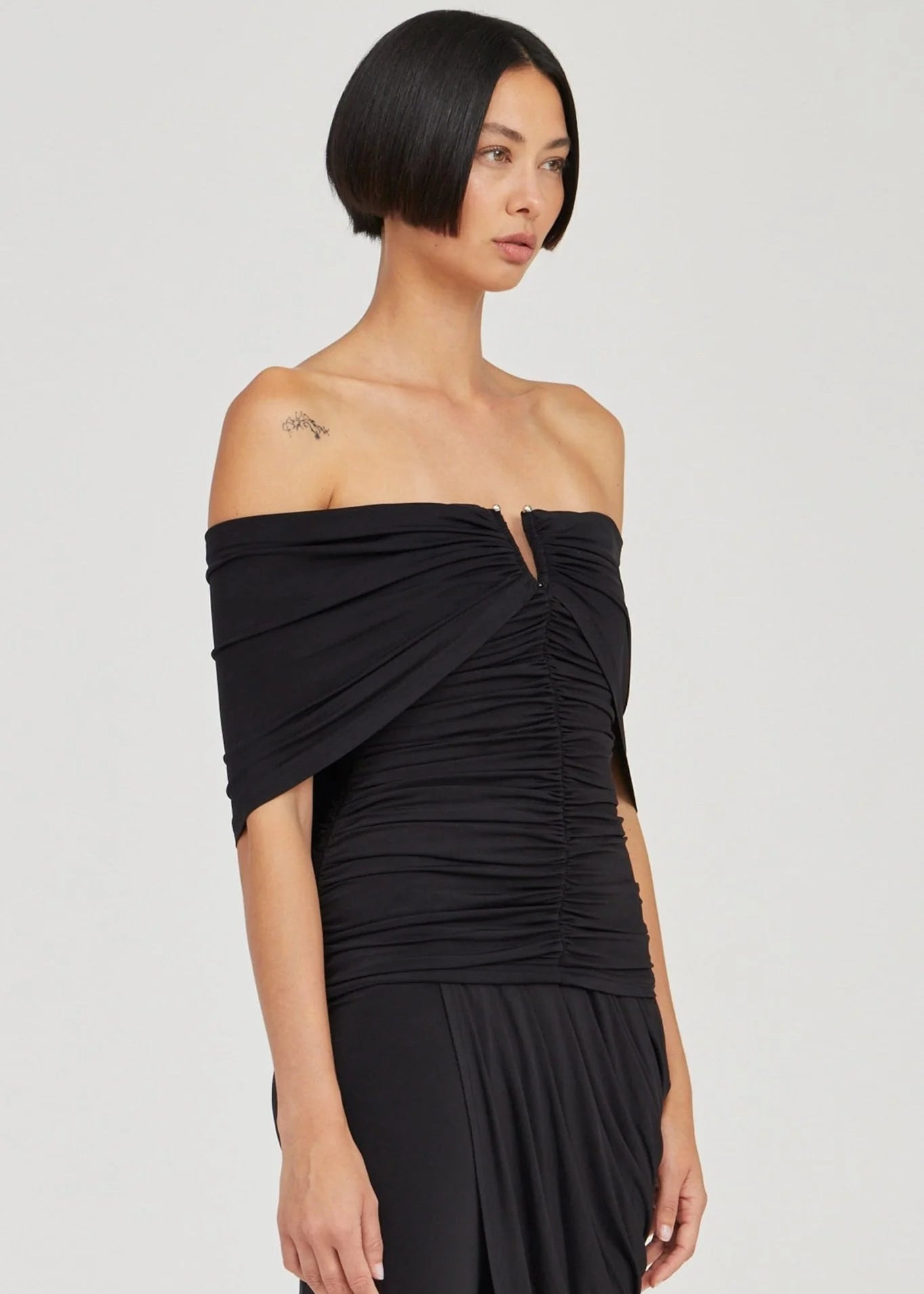 A woman modeling a black off-the-shoulder ruched dress with a split neckline and draped sleeve detail, photographed against a plain light gray studio background.