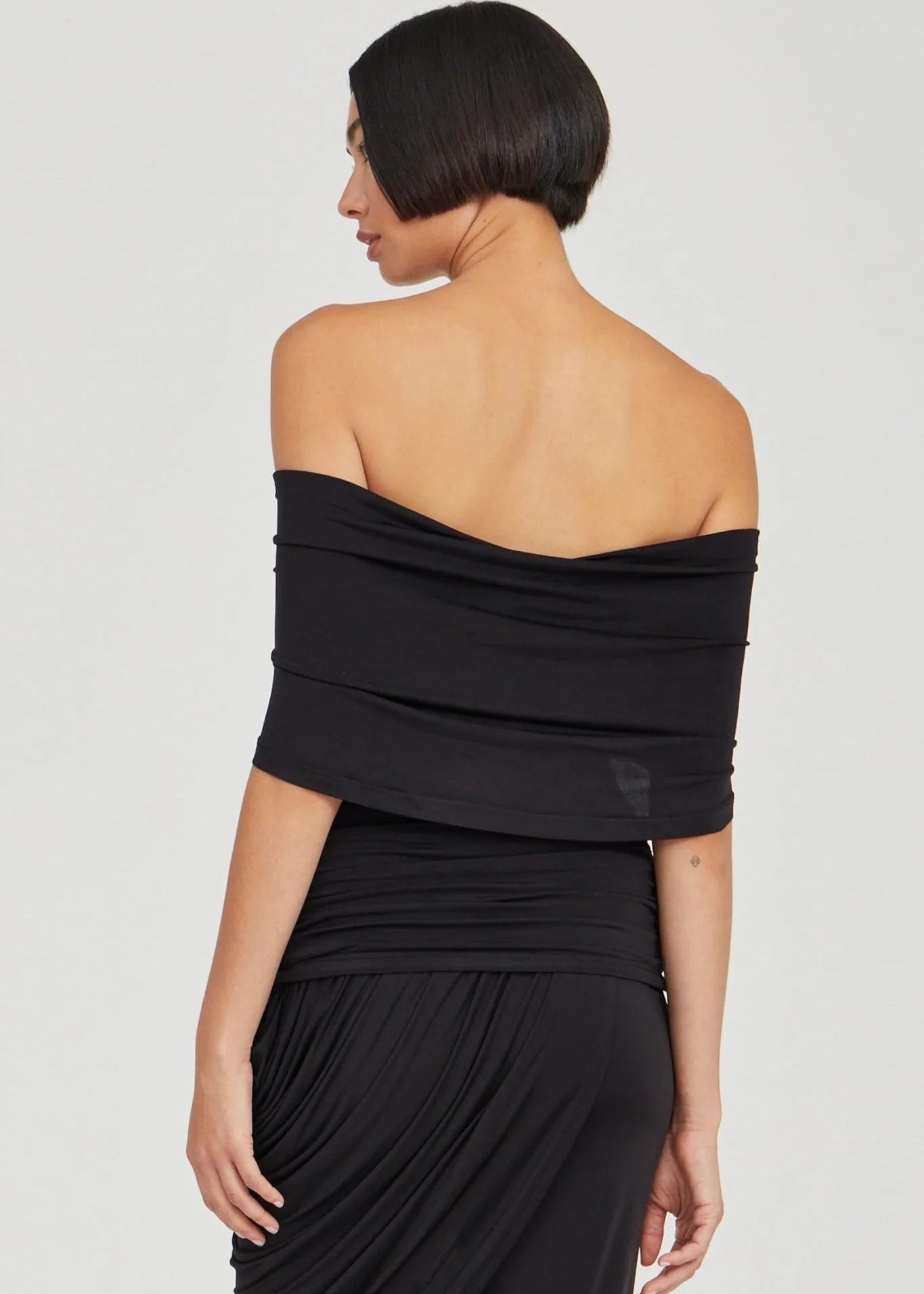 A woman shown from the back wearing a black off-the-shoulder draped top or dress with a gathered waist, photographed against a light gray studio background.