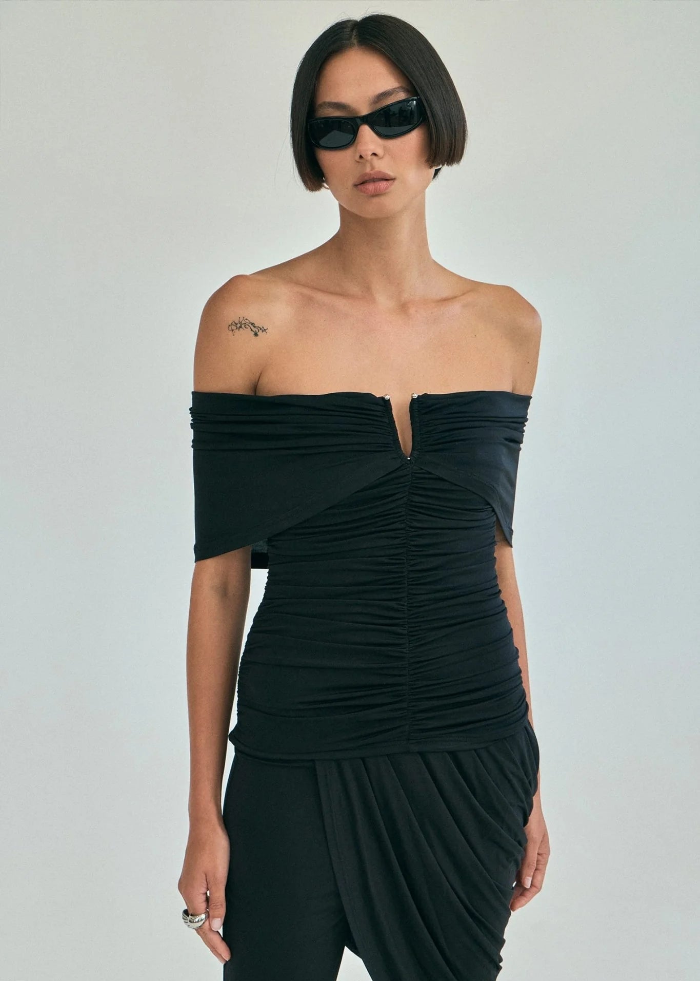 A model wearing a black off-the-shoulder ruched top and matching draped pants against a light gray studio background.