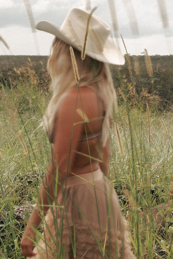 Person wearing a cowboy hat and brown outfit standing in a field with tall grass and wildflowers.
