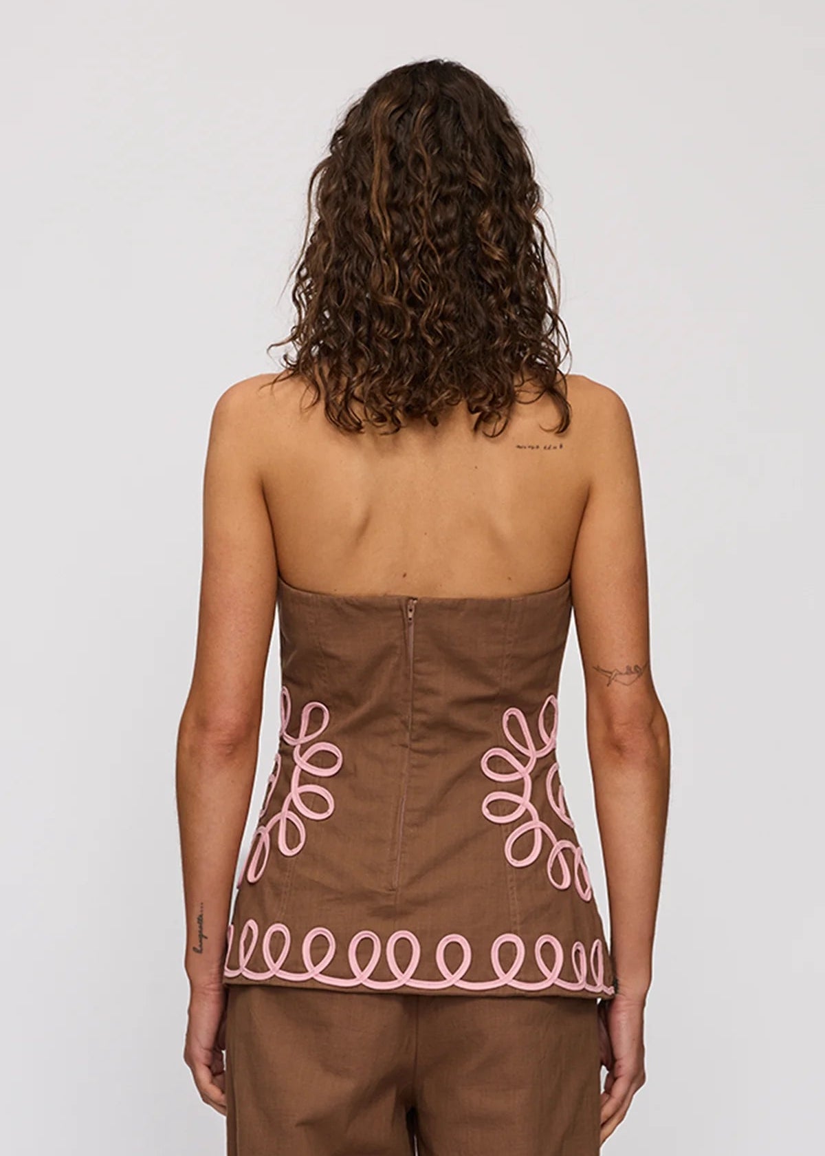 A woman viewed from behind wearing a brown strapless top with pink embroidered floral and wave patterns, paired with matching brown pants, against a light gray studio background.