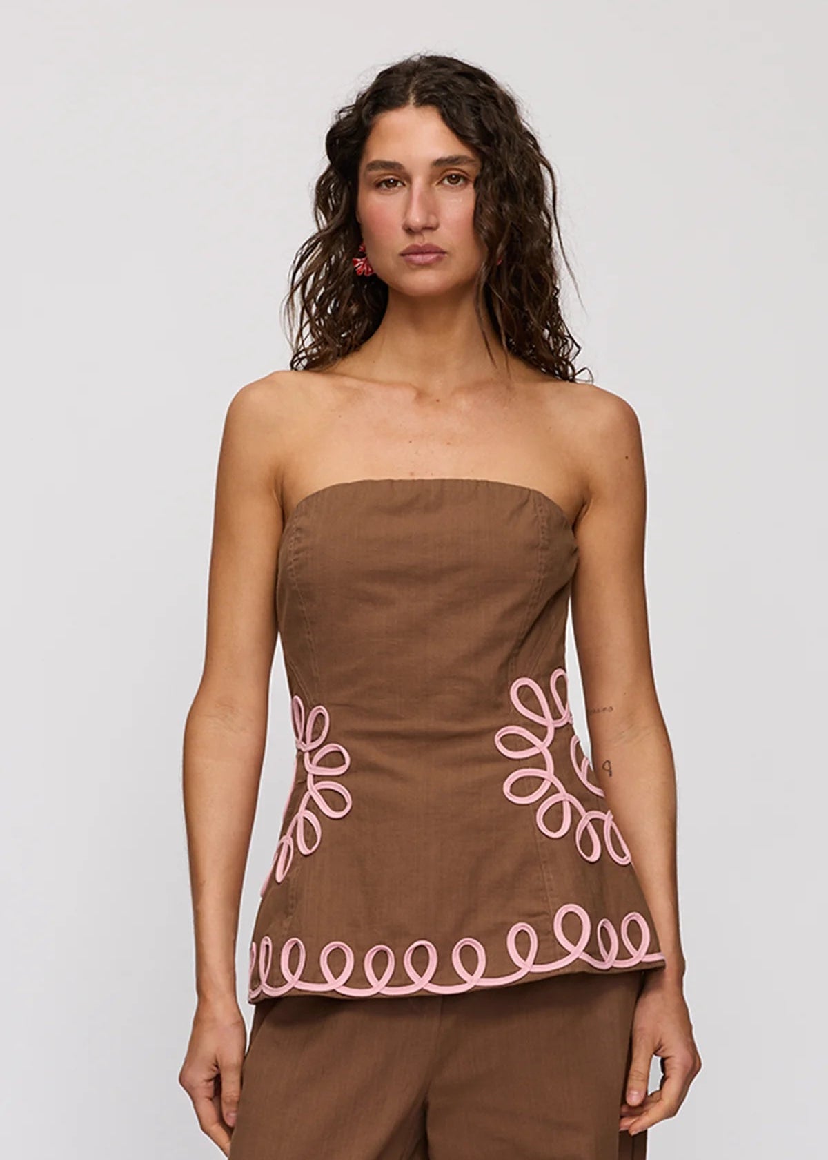 A woman modeling a brown strapless top with pink embroidered floral and scalloped details, paired with matching brown pants, against a light gray studio background.