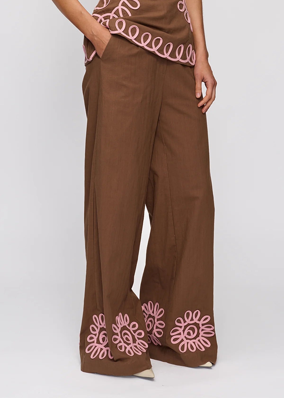 Brown wide-leg trousers with pink floral embroidery at the hem, shown on a model against a light gray background.