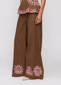 A model wearing brown wide-leg pants with pink embroidered floral patterns near the hems, paired with a matching brown top, shot against a light gray studio background.