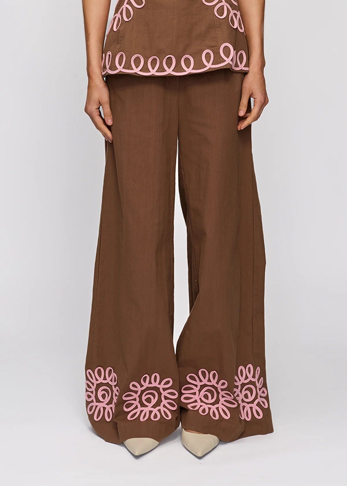 A person wearing brown wide-leg trousers with pink embroidered floral and scalloped details near the hem, paired with a matching brown top, shown against a light gray studio background.