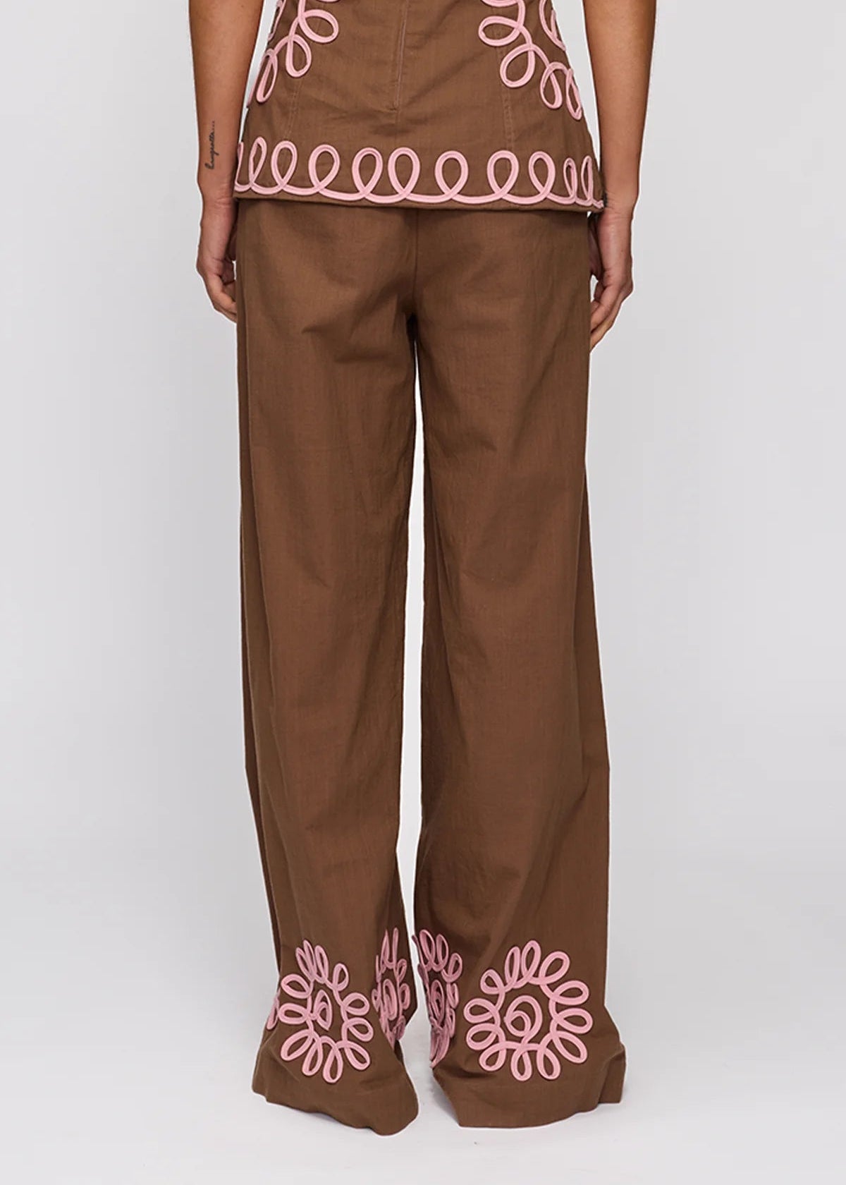 A person shown from the back wearing brown wide-leg pants with pink floral embroidery near the hem and a matching brown top with pink circular embroidery near the waist, against a light gray studio background.