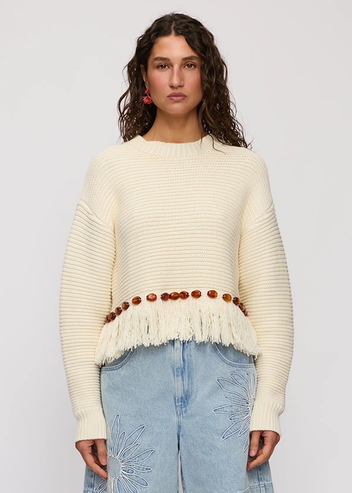 A woman modeling a cream ribbed knit sweater with a fringed hem and a brown beaded belt, paired with light blue jeans, against a light gray studio background.