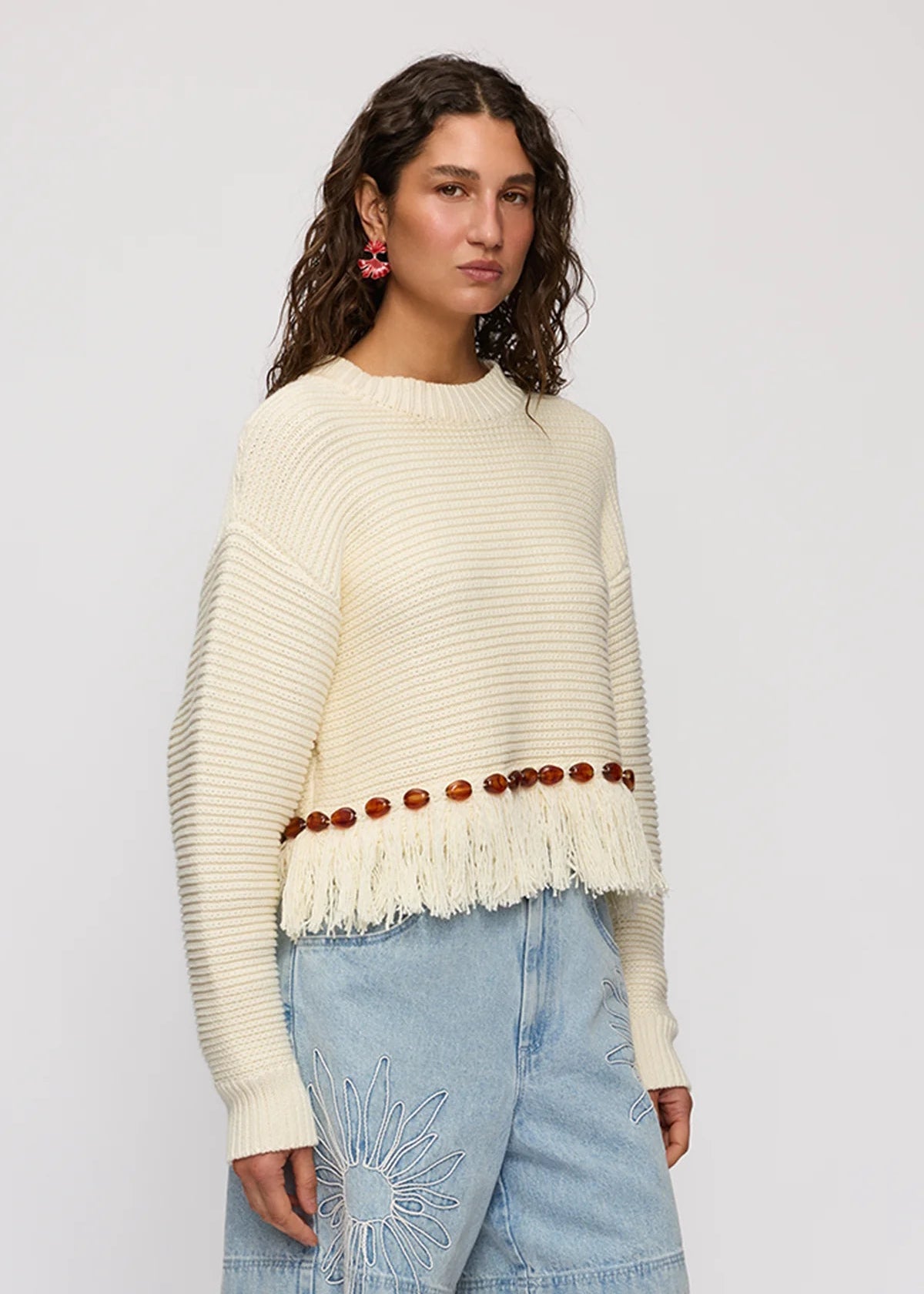 A woman modeling a cream ribbed knit sweater with a fringed hem and a row of brown wooden beads, paired with light blue denim shorts against a light gray studio background.
