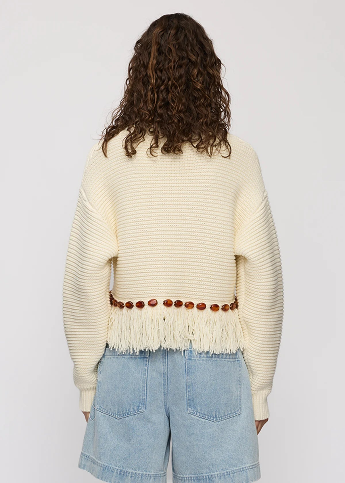 A model viewed from behind wearing a cream-colored ribbed knit sweater with fringe and a brown beaded waist belt, paired with light blue denim shorts, against a plain light gray background.