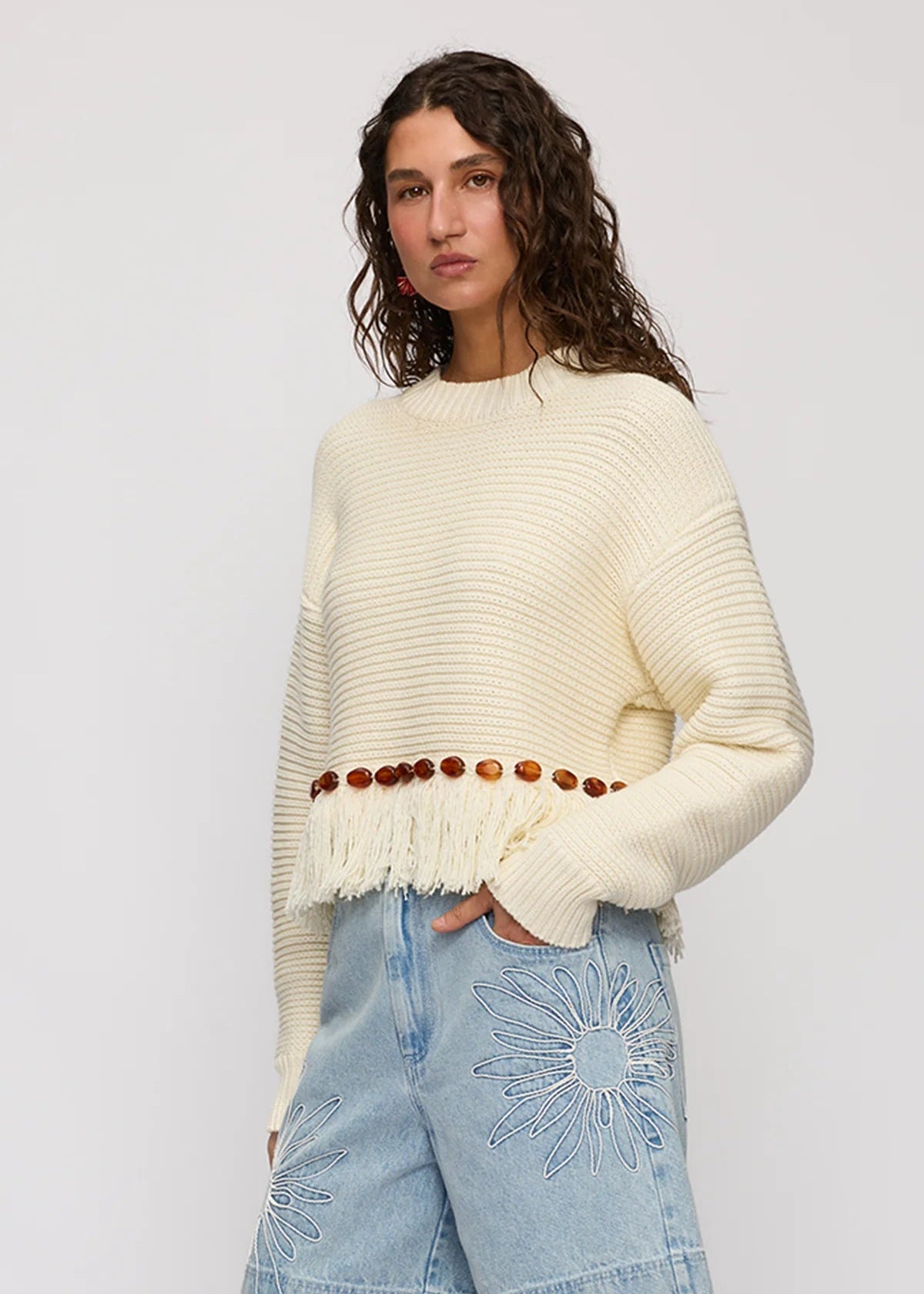 A woman wearing a cream ribbed knit sweater with fringe and a brown beaded waist detail, paired with light blue denim shorts, against a light gray studio background.