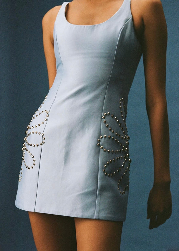 A sleeveless light blue mini dress with metallic beaded decorative details on the sides, shown on a model against a dark blue background.