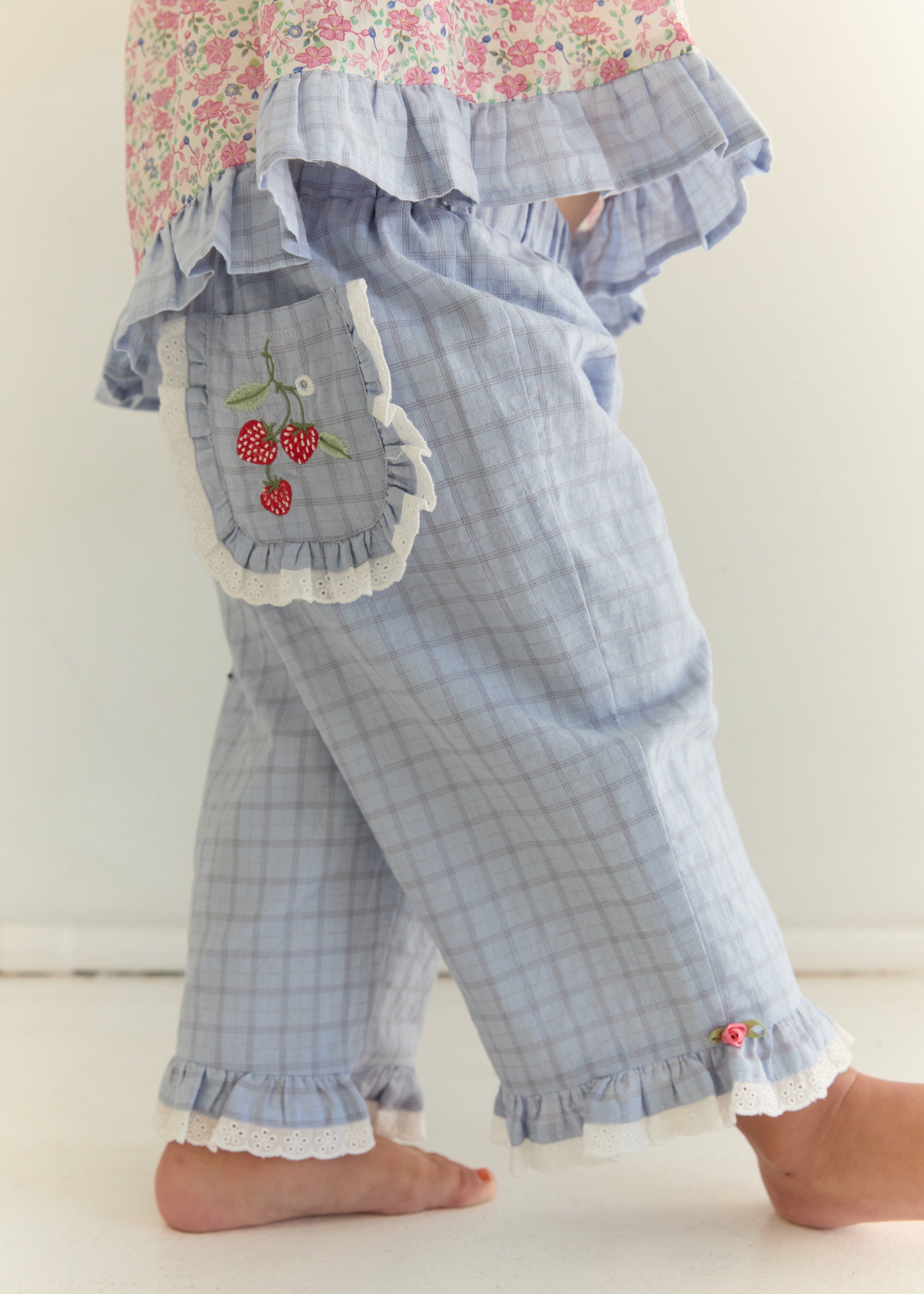 A close-up of a child's legs wearing light blue plaid ruffled pants with a cherry embroidered pocket and white lace trim, paired with a floral top.