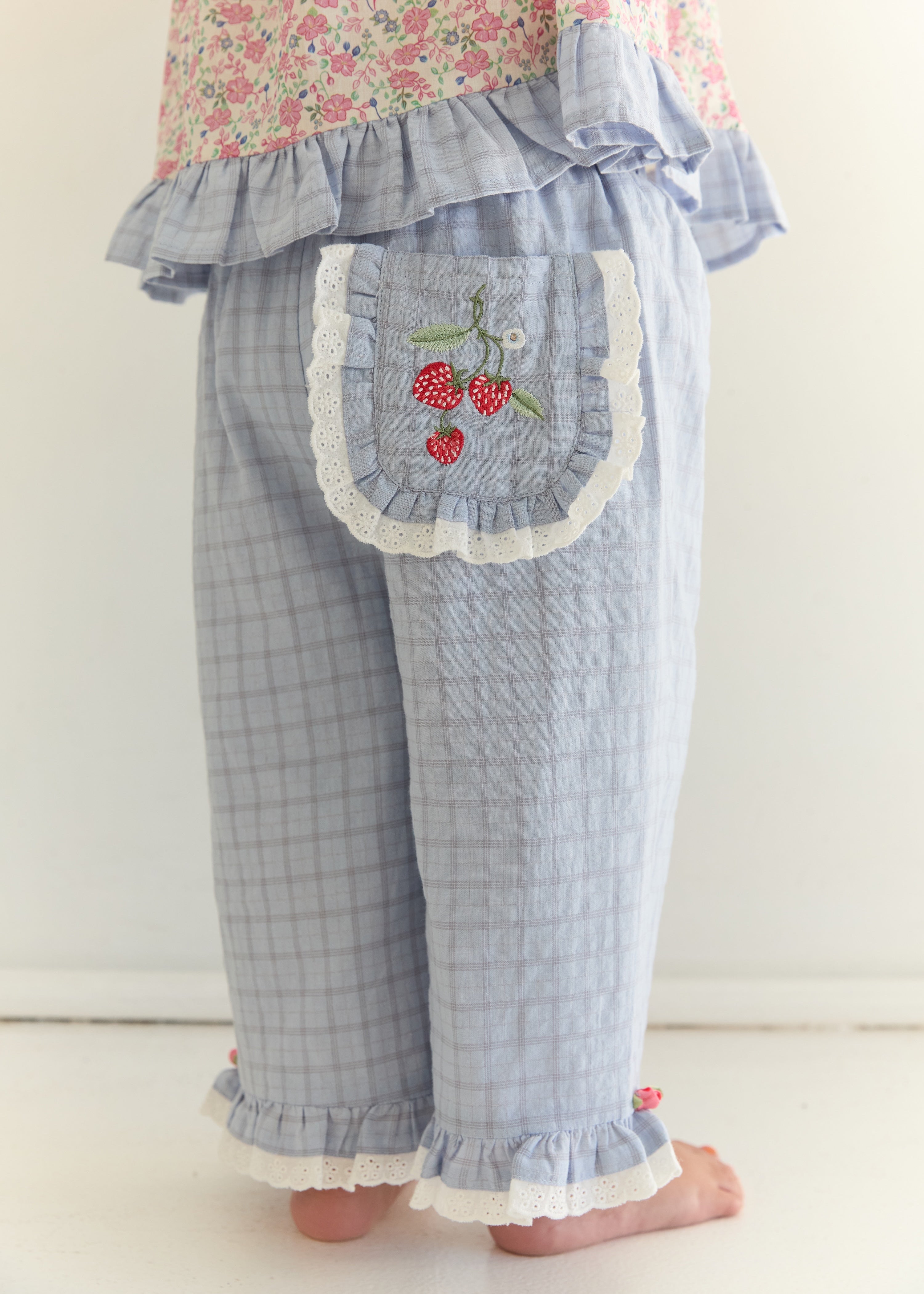A child's light blue plaid pants with a decorative back pocket featuring white lace trim and embroidered red cherries, shown from the waist down against a plain white background.