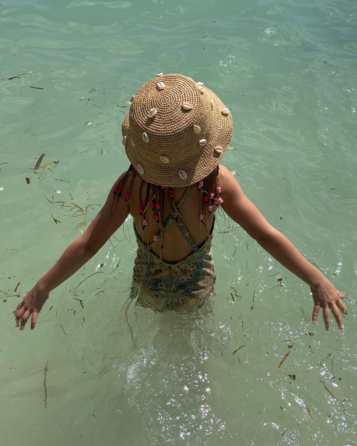 A child standing in shallow, clear turquoise water, viewed from behind, wearing a woven straw sun hat decorated with small seashells and a patterned swimsuit.