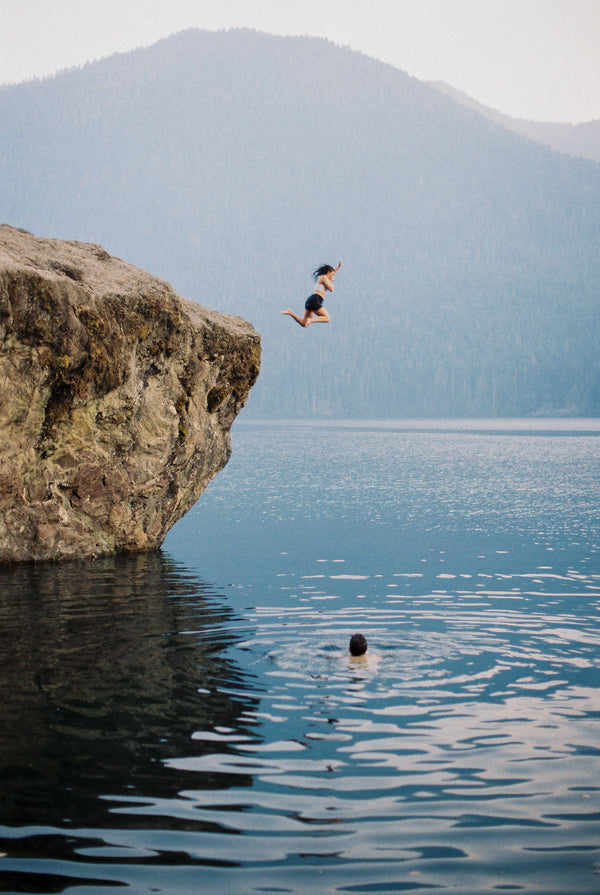 Person jumping off a cliff into a body of water with mountains in the background