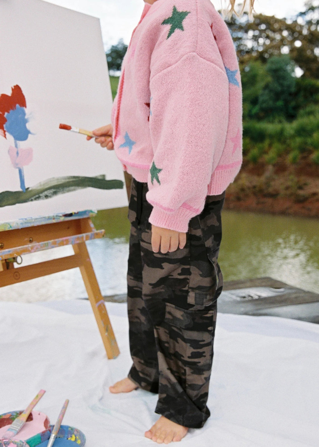 Child in pink sweater and camouflage pants painting on an easel outdoors.