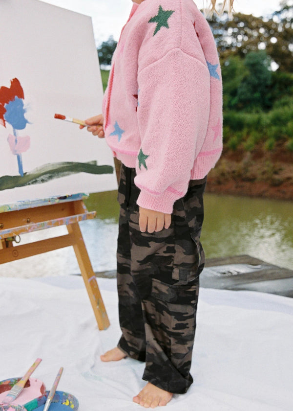 Child in pink sweater and camouflage pants painting on an easel outdoors.