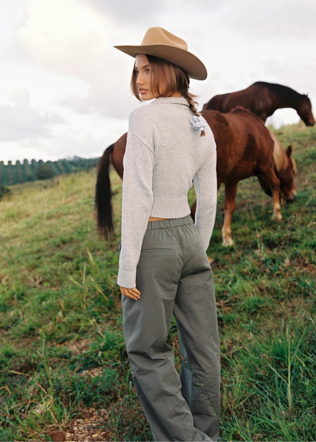 Woman in a field with horses wearing a beige hat and light gray sweater.